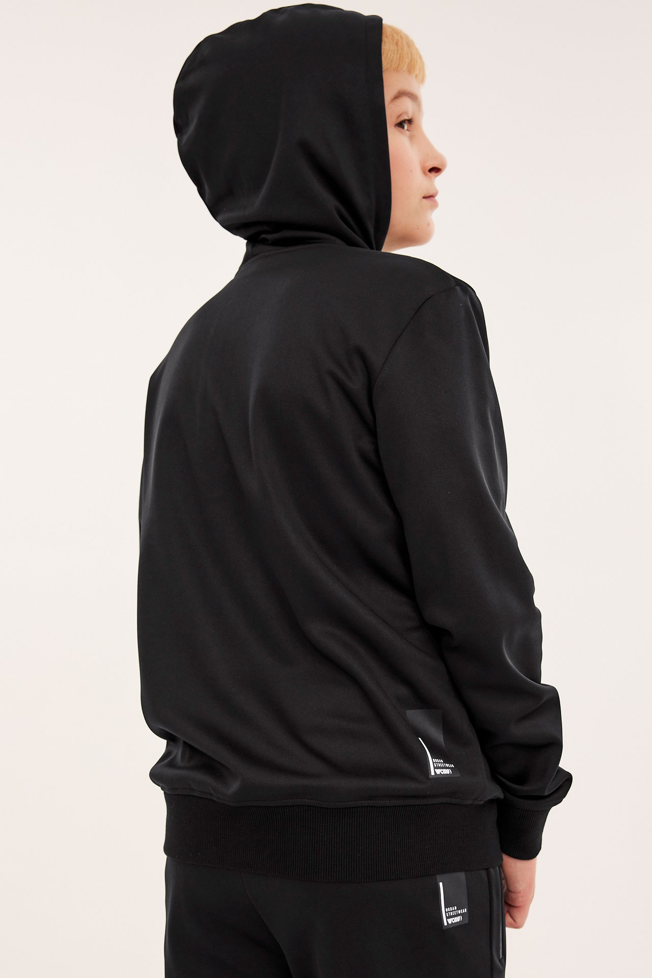 Sidevisning av et barn i en svart ensfarget polyesterhoodie med hette oppe og matchende joggebukse.