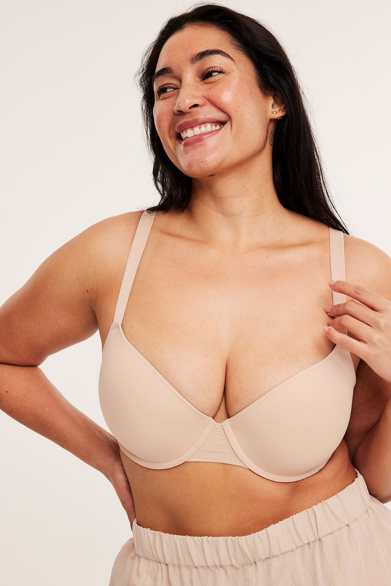 Forfra, smilende kvinne i en lys beige glatt push-up BH og lys beige shorts med elastisk midje.