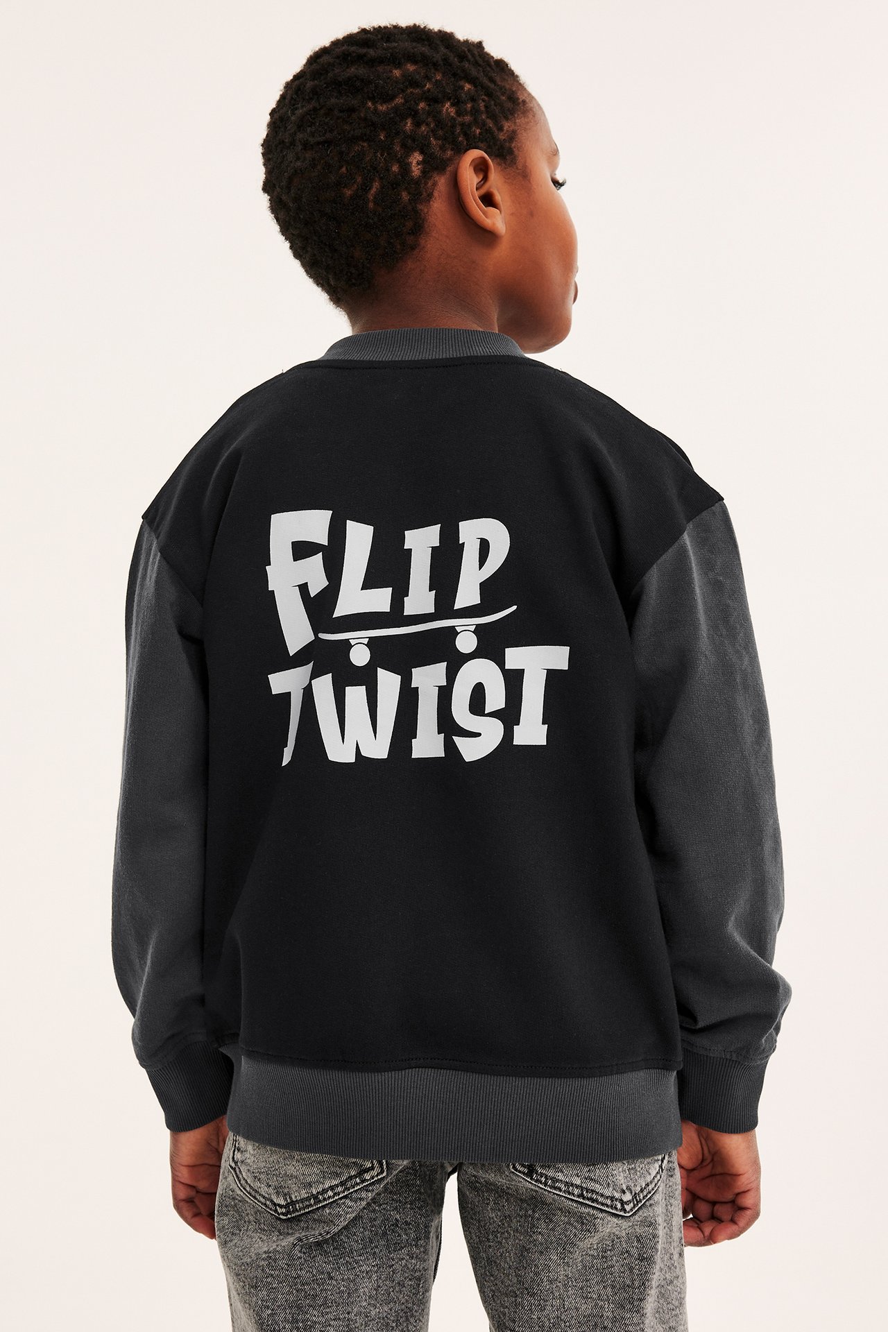 Bakfra: et barn i svart og grå baseballjakke med 'FLIP TWIST'-trykk og grå jeans.