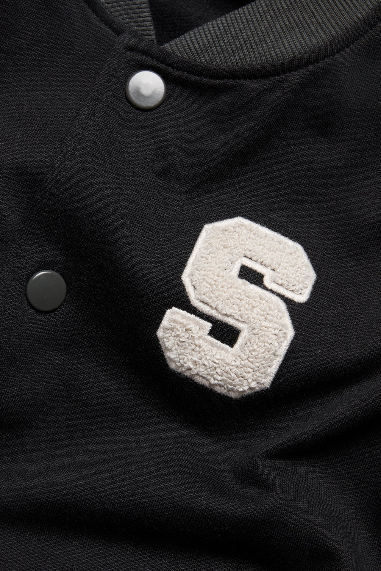 Nærbilde forfra av en svart bomulls baseballjakke med hvit frotté 'S'-applikasjon og trykknapper.