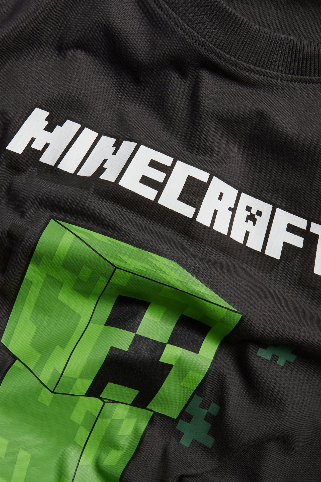 Nærbilde av en svart bomulls-T-skjorte for barn med stort grønt Minecraft Creeper-trykk og hvit "MINECRAFT"-tekst.