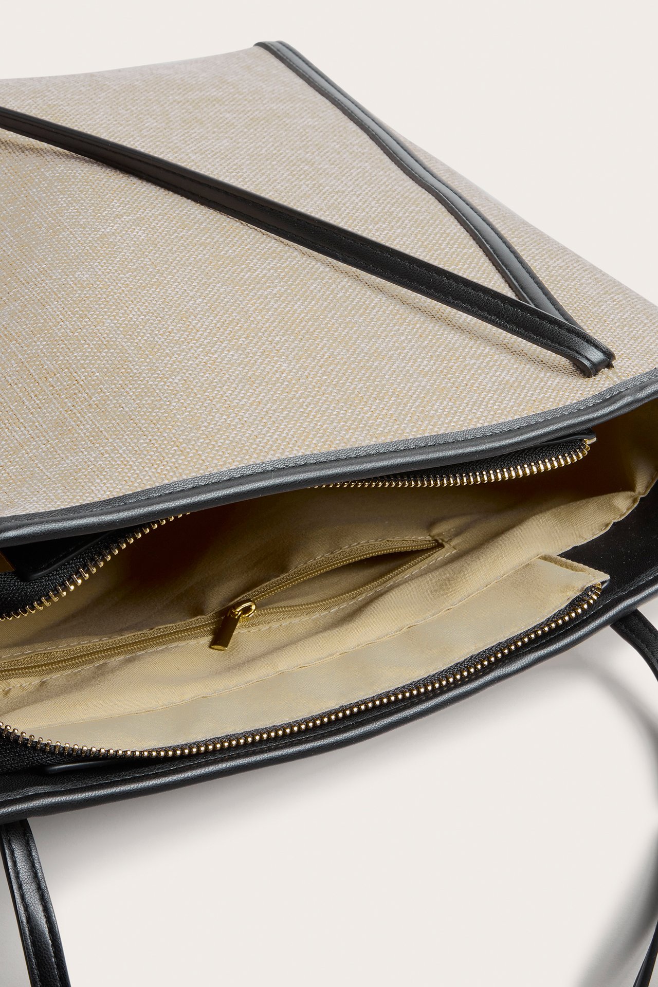 Närbild, uppifrån, av en öppen beige strå-look tygväska med svarta läderimitation detaljer och innerficka med dragkedja.