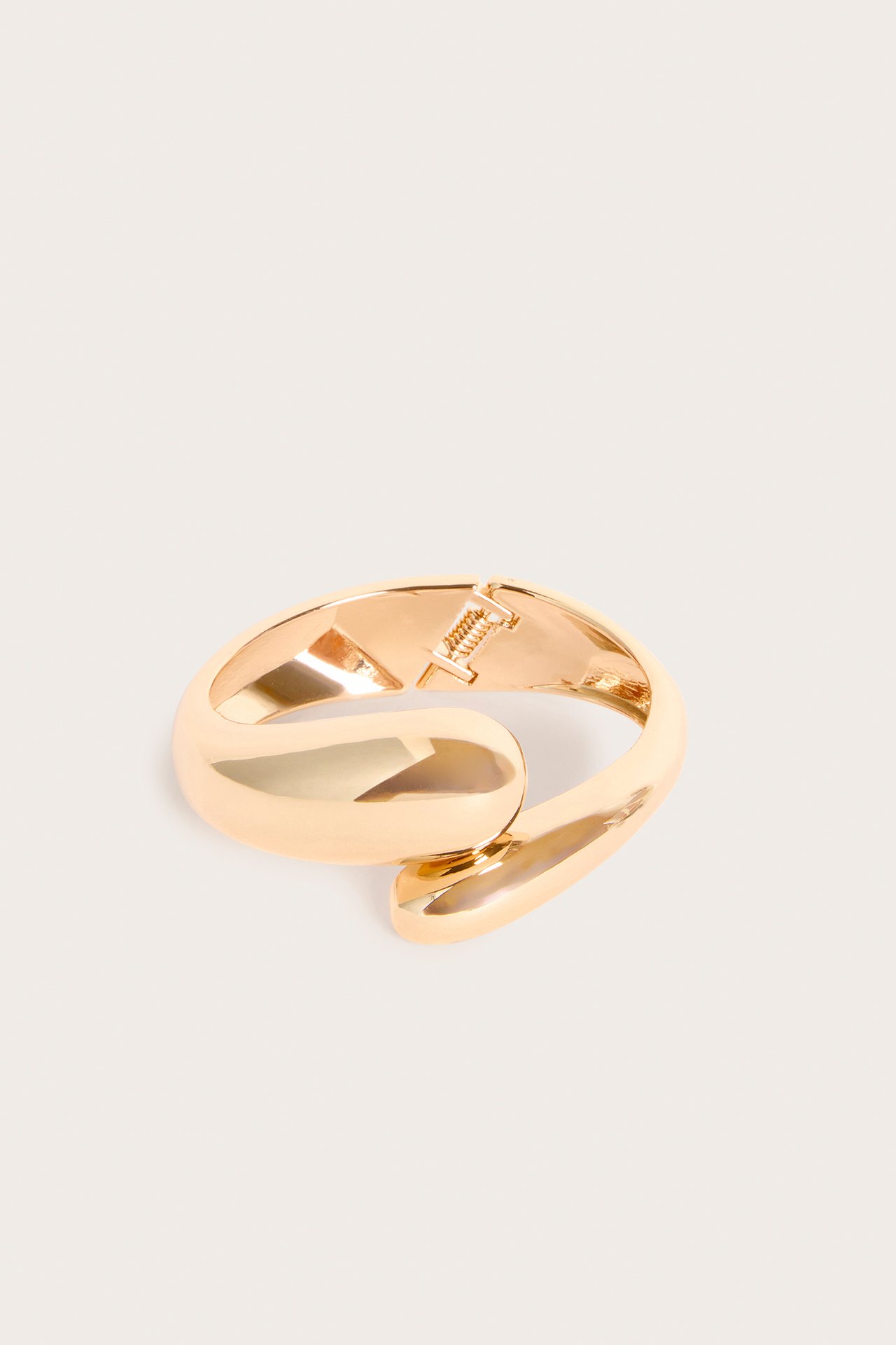Nærbilde av et gullfarget stivt armbånd med bølget, skulpturell form og hengselfeste.