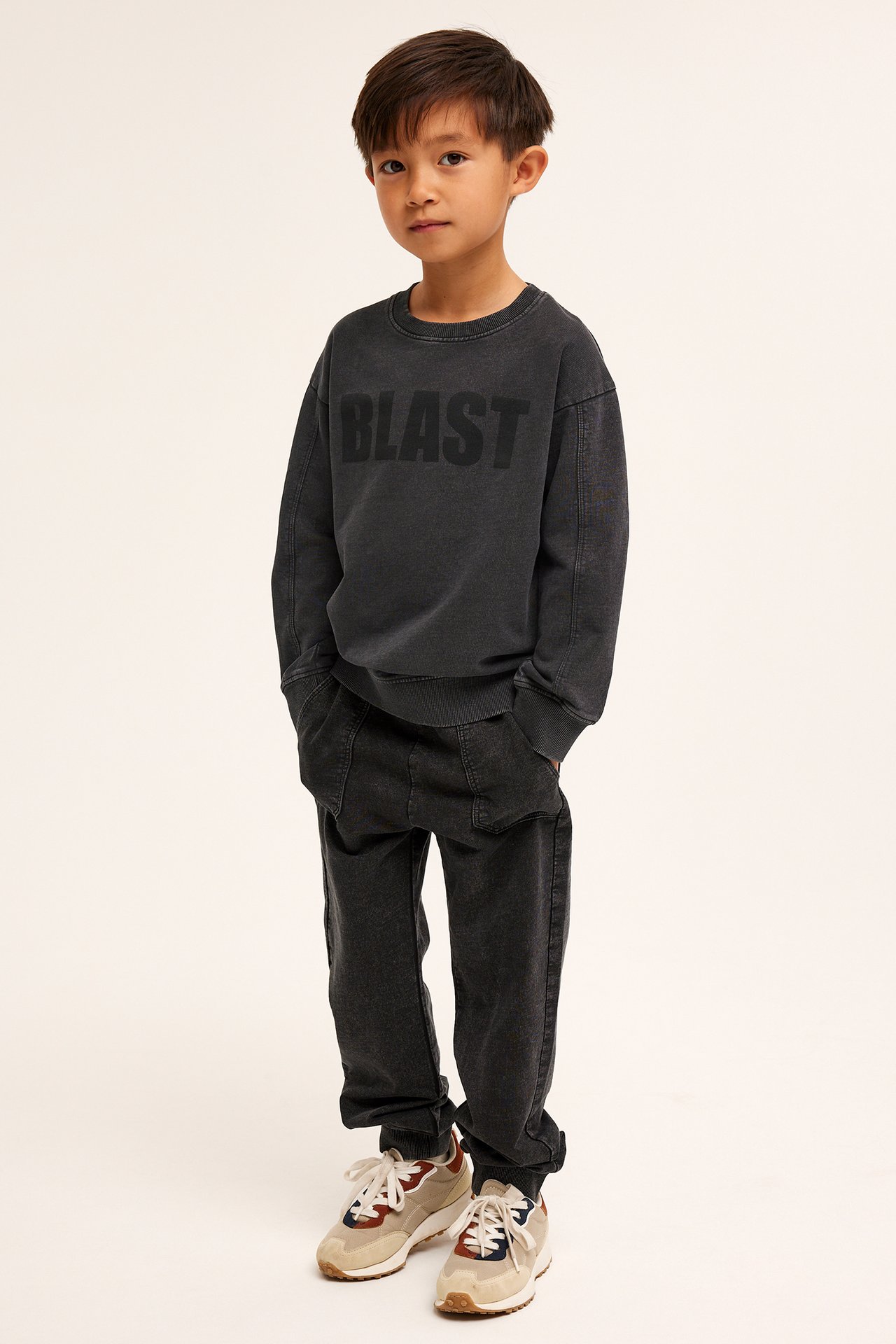 Frontvy av ett barn i en mörkgrå tvättad bomullssweatshirt med 'BLAST'-tryck, matchande joggingbyxor och sneakers.