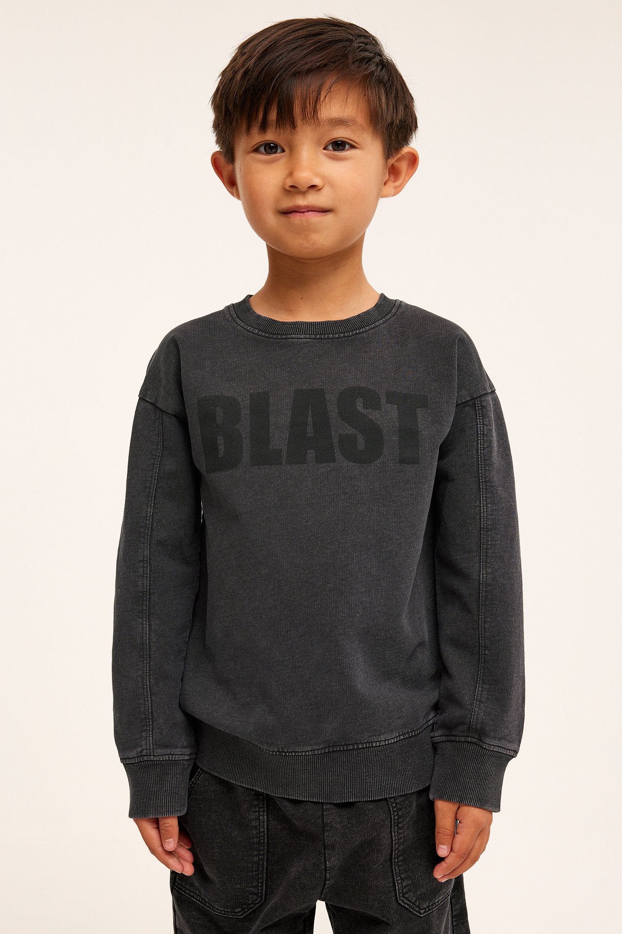 Frontvy av ett barn i en svart tvättad bomullssweatshirt med "BLAST"-tryck och svarta byxor.