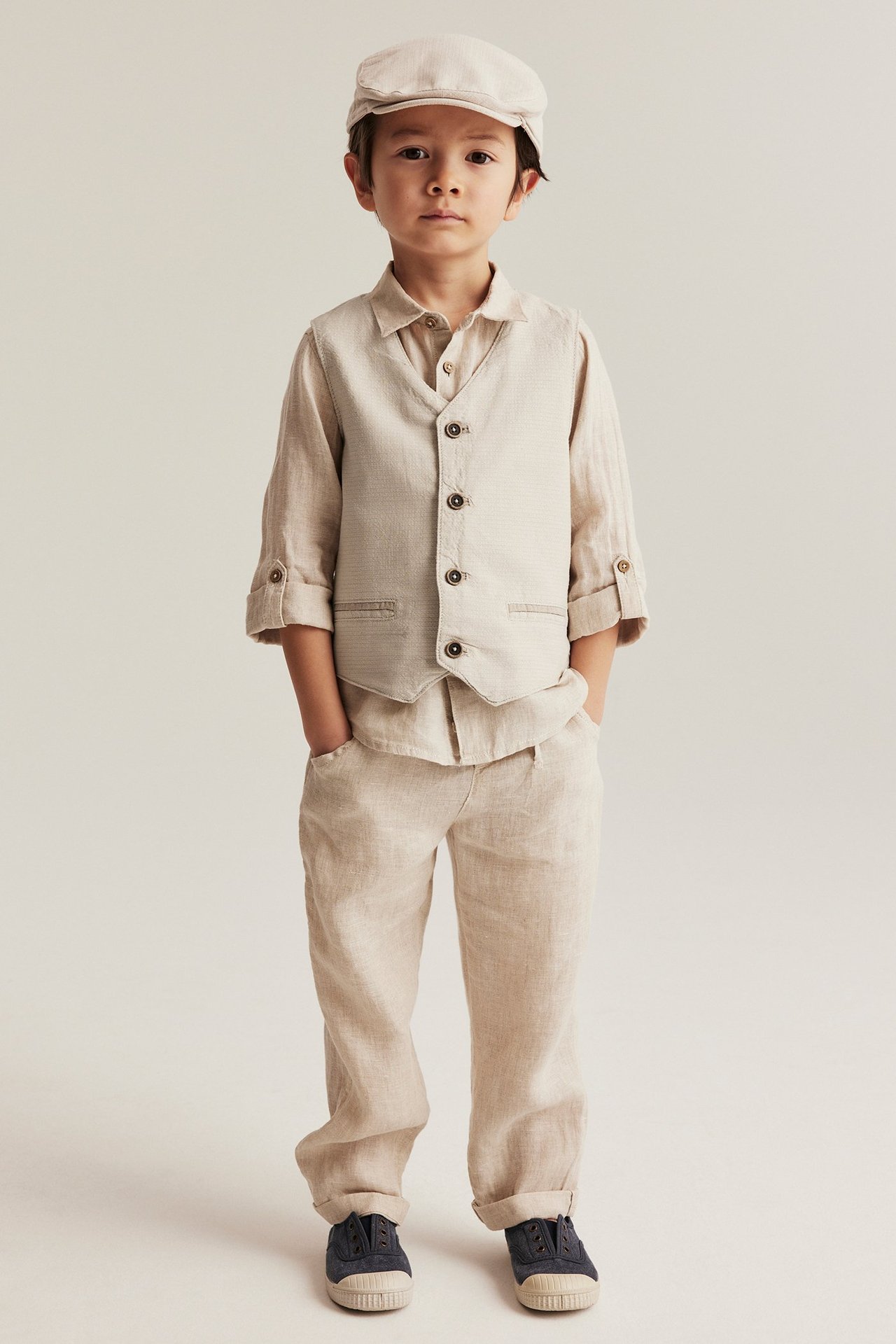 Framifrån: Kid i beige vävd väst med knappar, beige skjorta, matchande byxor, beige keps och blå skor.