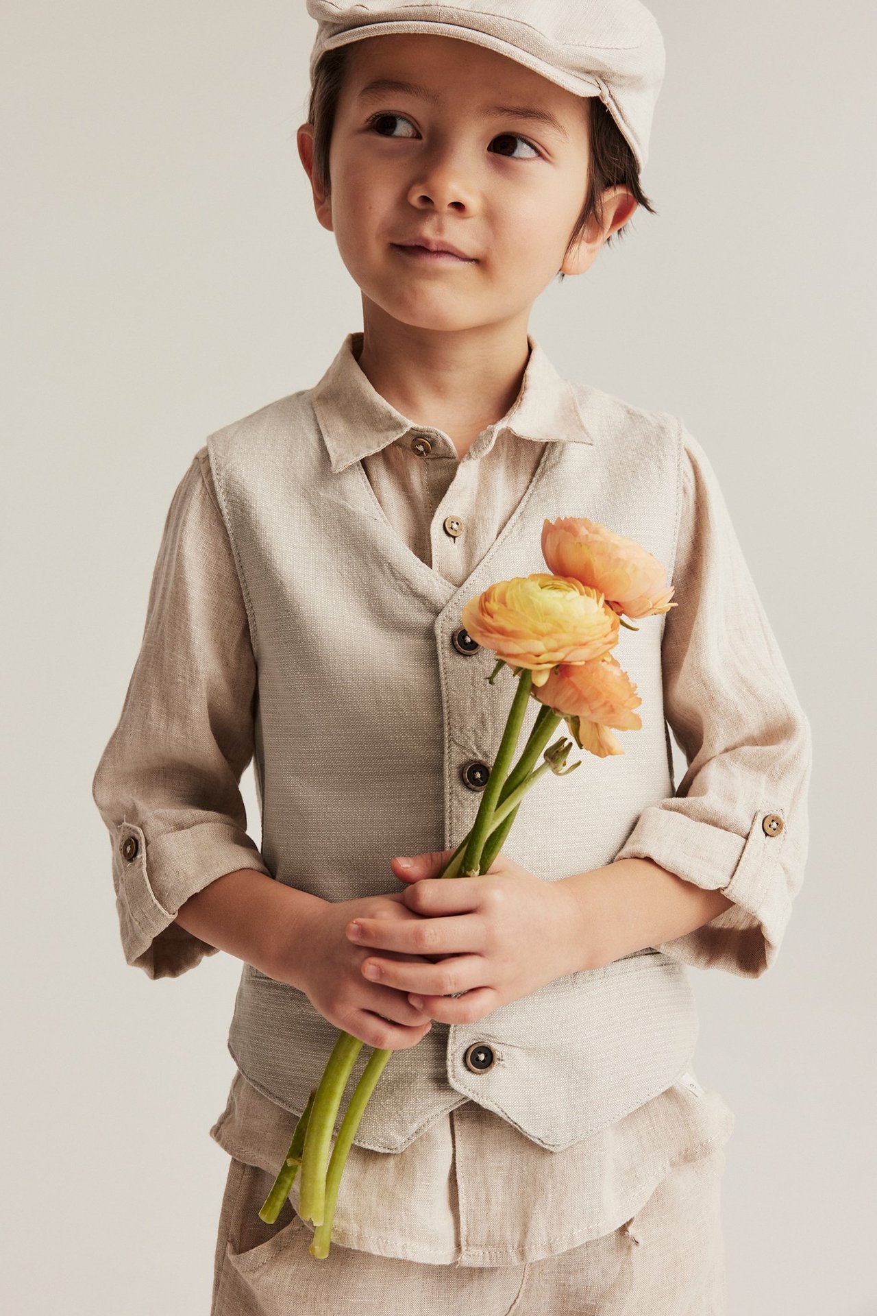 Barn i beige vävd väst, matchande skjorta, byxor och keps, håller orange blommor.