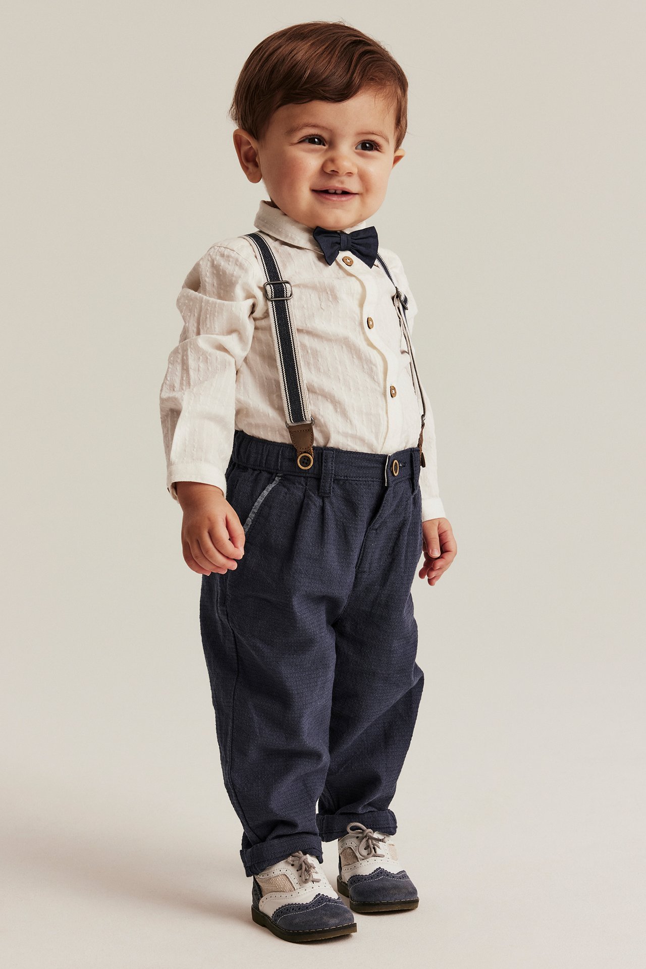 Edestä: vauva tummansinisissä kudotuissa housuissa henkseleillä, vaalea paita, tummansininen rusetti.