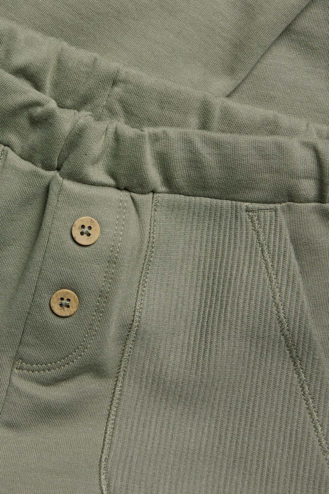 Zbliżenie na ciemnozielone bawełniane spodnie dresowe z elastyczną talią, ozdobnymi guzikami i prążkowaną kieszenią.