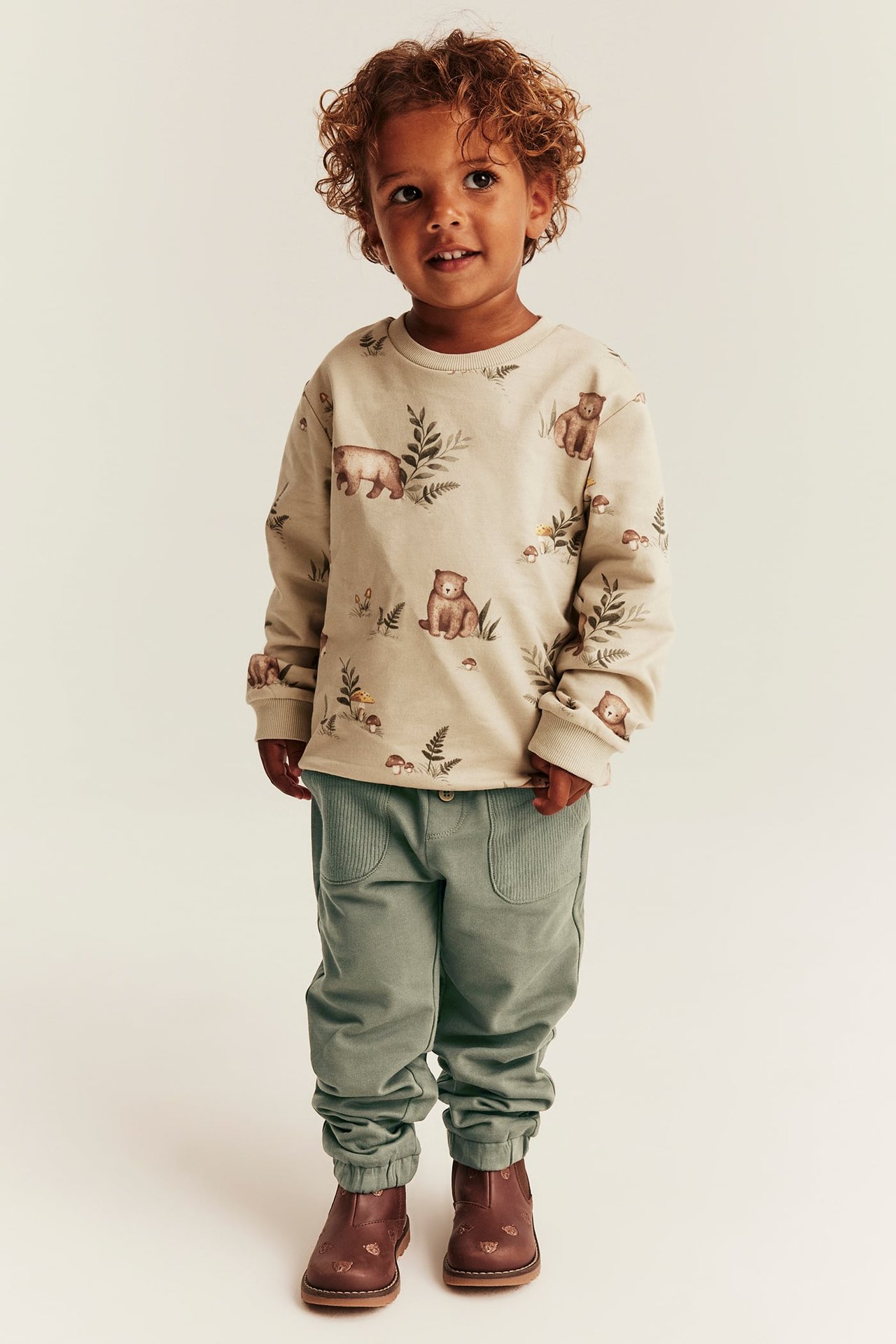 Frontvisning barnas langermet beige topp med bjørnetrykk, salviegrønne joggebukser og brune ankelstøvler.
