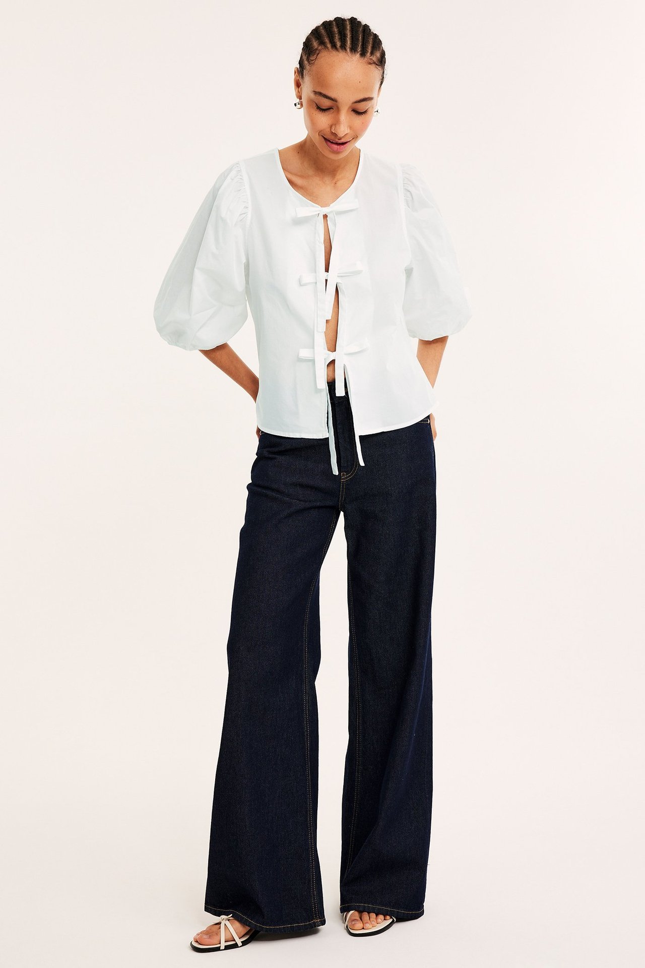 Framifrån: kvinna i vit bomullsblus med puffärm och knytband, mörkblå vida jeans, vita sandaler.