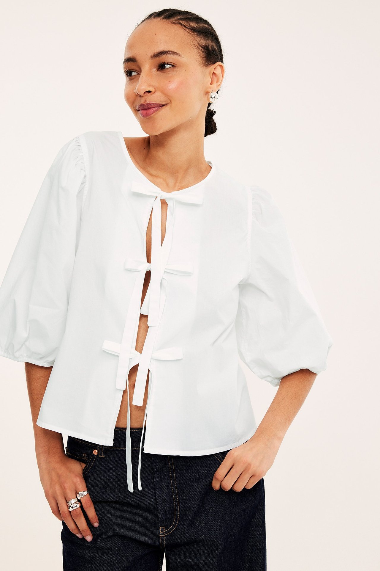 Widok z przodu: kobieta w białej bawełnianej bluzce z bufiastymi rękawami, wiązaniami, ciemnoniebieskich dżinsach, srebrnych pierścionkach, perłowych kolczykach.