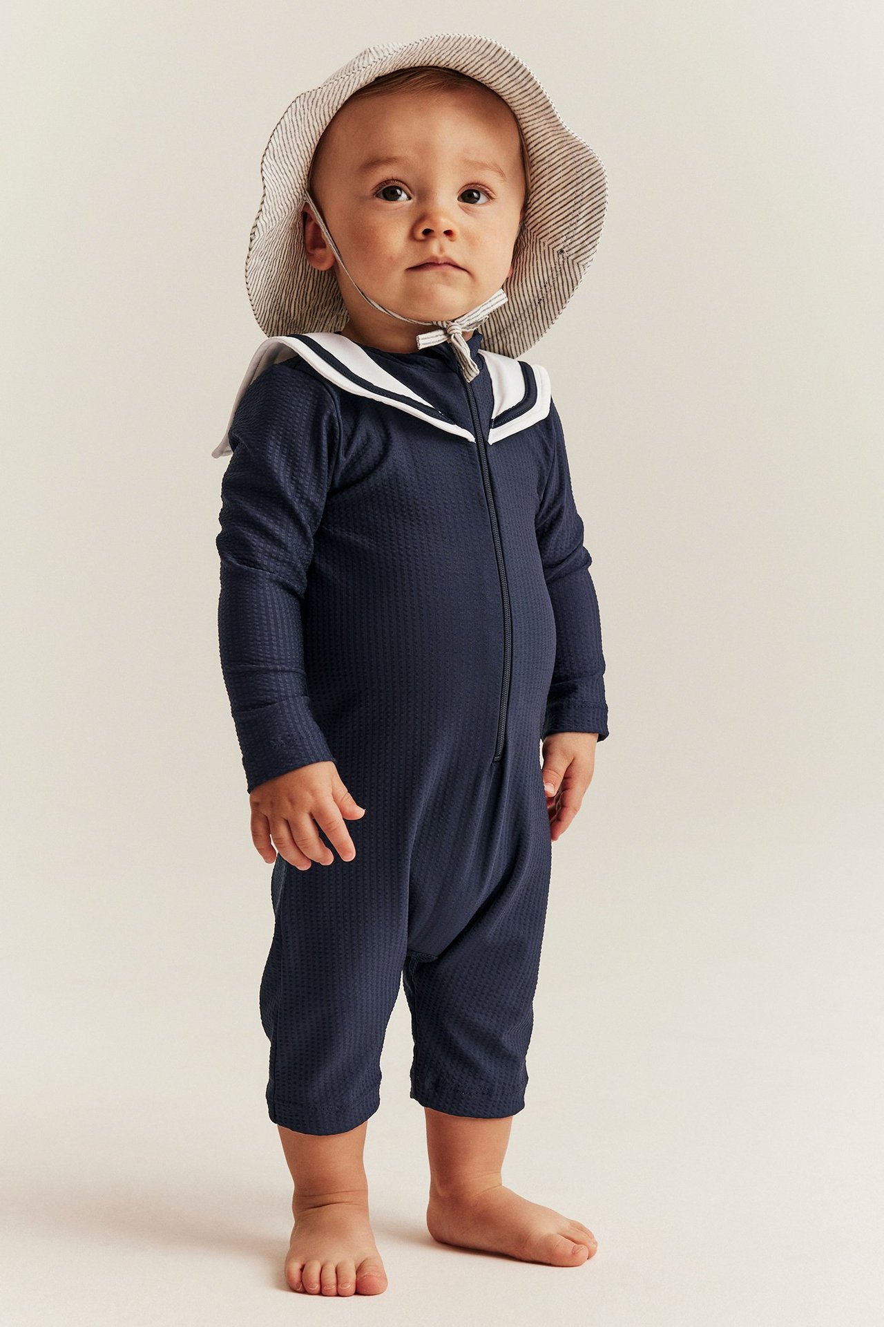Frontvy av baby i mörkblå UV-dräkt med vit sjömanskrage och randig solhatt.