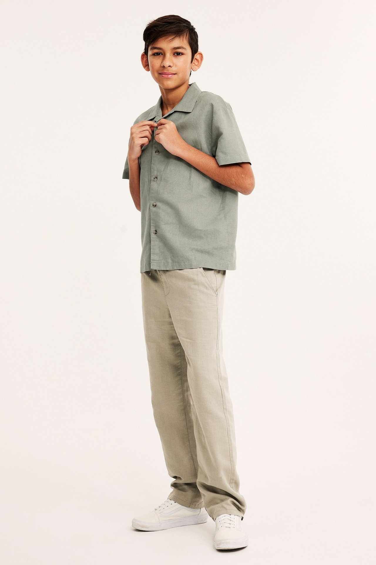 Widok z boku dziecka w beżowych spodniach z mieszanki lnu, zielonkawo-szarej koszuli i białych trampkach.