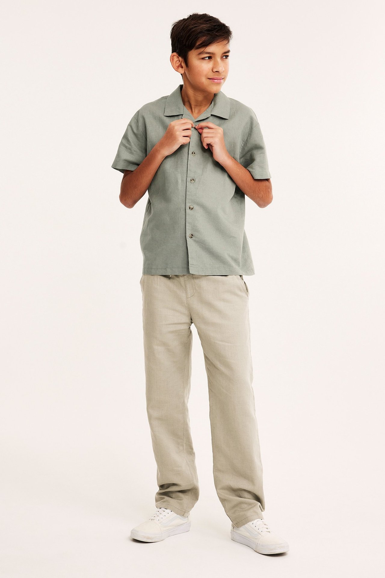 Widok z przodu dziecka w beżowych spodniach z mieszanki lnu, jasnozielonej koszuli i białych trampkach.