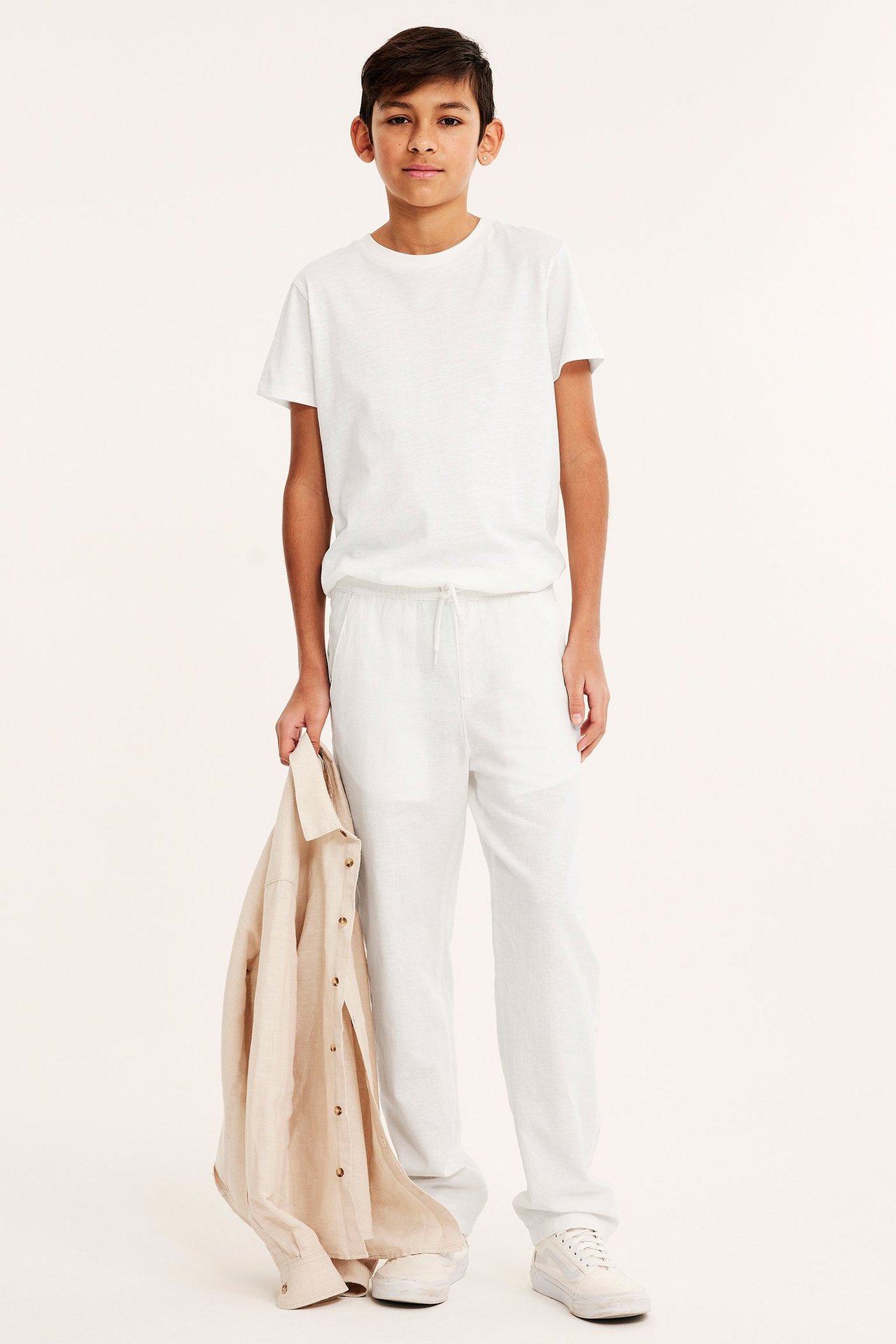 Widok z przodu: Dziecko w ecru lnianych spodniach, białym t-shircie, białych sneakersach, trzyma beżową koszulę.