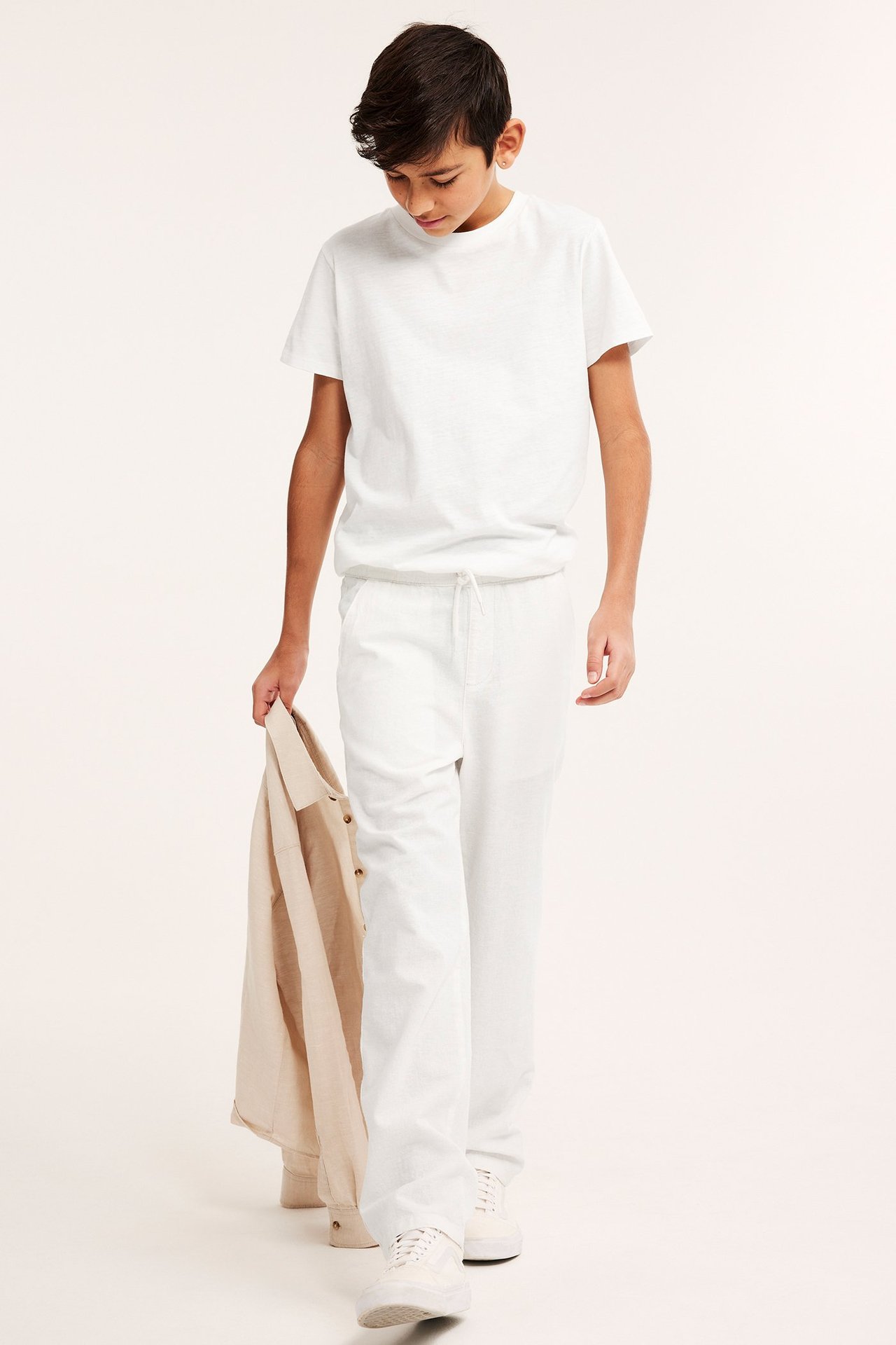Widok z przodu: Dziecko w luźnych, ecru spodniach lnianych ze sznurkiem, białym T-shircie, trzyma beżową koszulę.