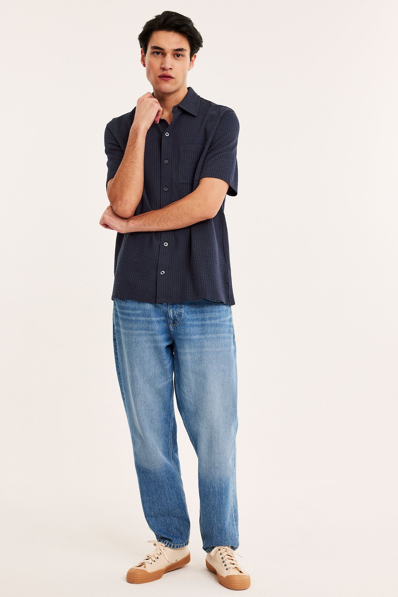 Edestäpäin mies tummansinisessä kuvioidussa lyhythihaisessa neulepaidassa, vaaleansinisissä farkuissa ja beigenvärisissä tennareissa.