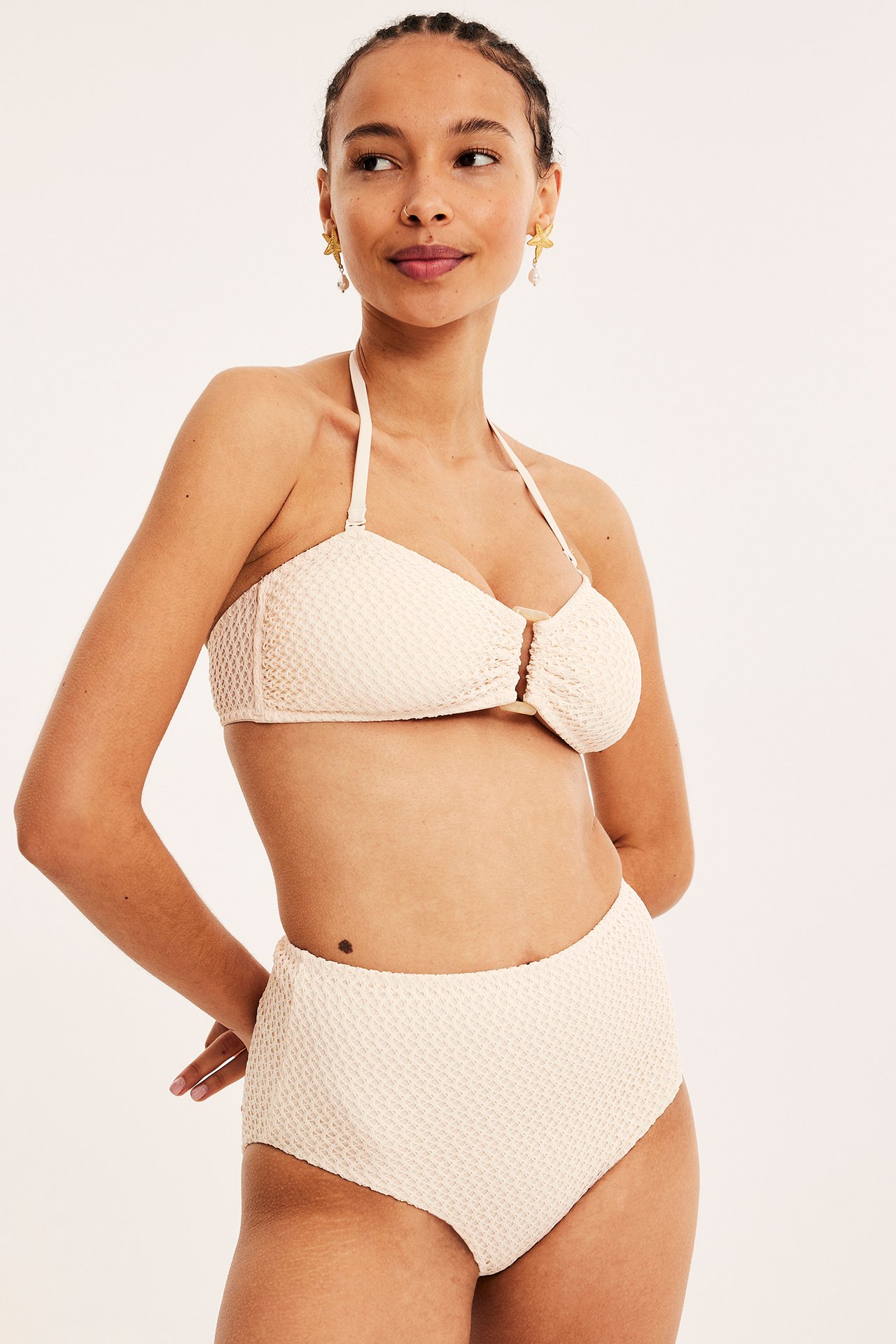 Framifrån: kvinna i vita texturerade högmidjade bikinitrosor, matchande bandeau-topp och guldiga sjöstjärneörhängen.