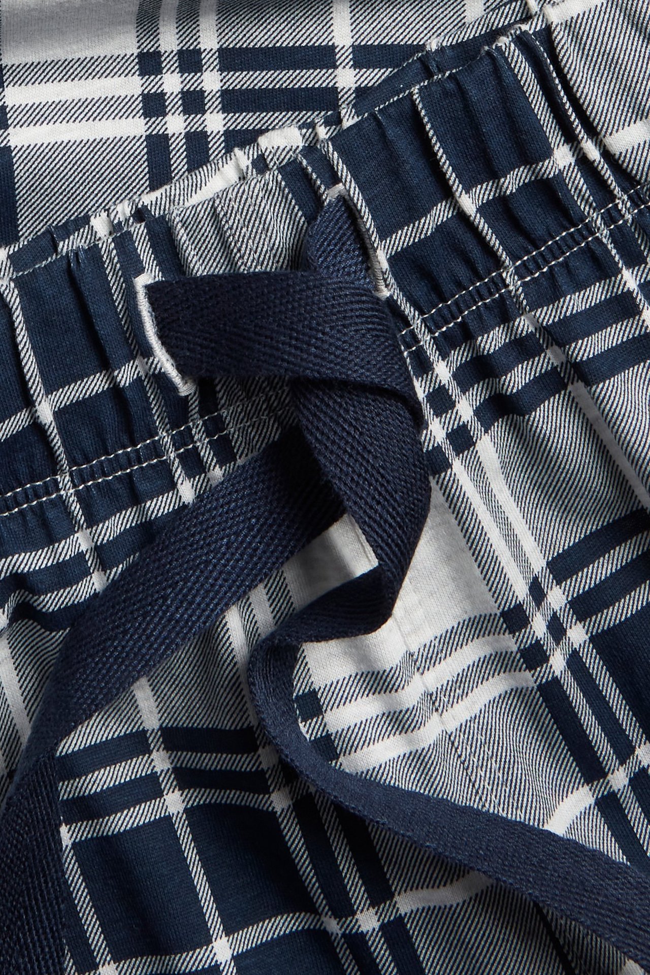 Zbliżenie na granatowe i białe piżamowe spodnie w kratę z elastyczną talią i granatowym sznurkiem.