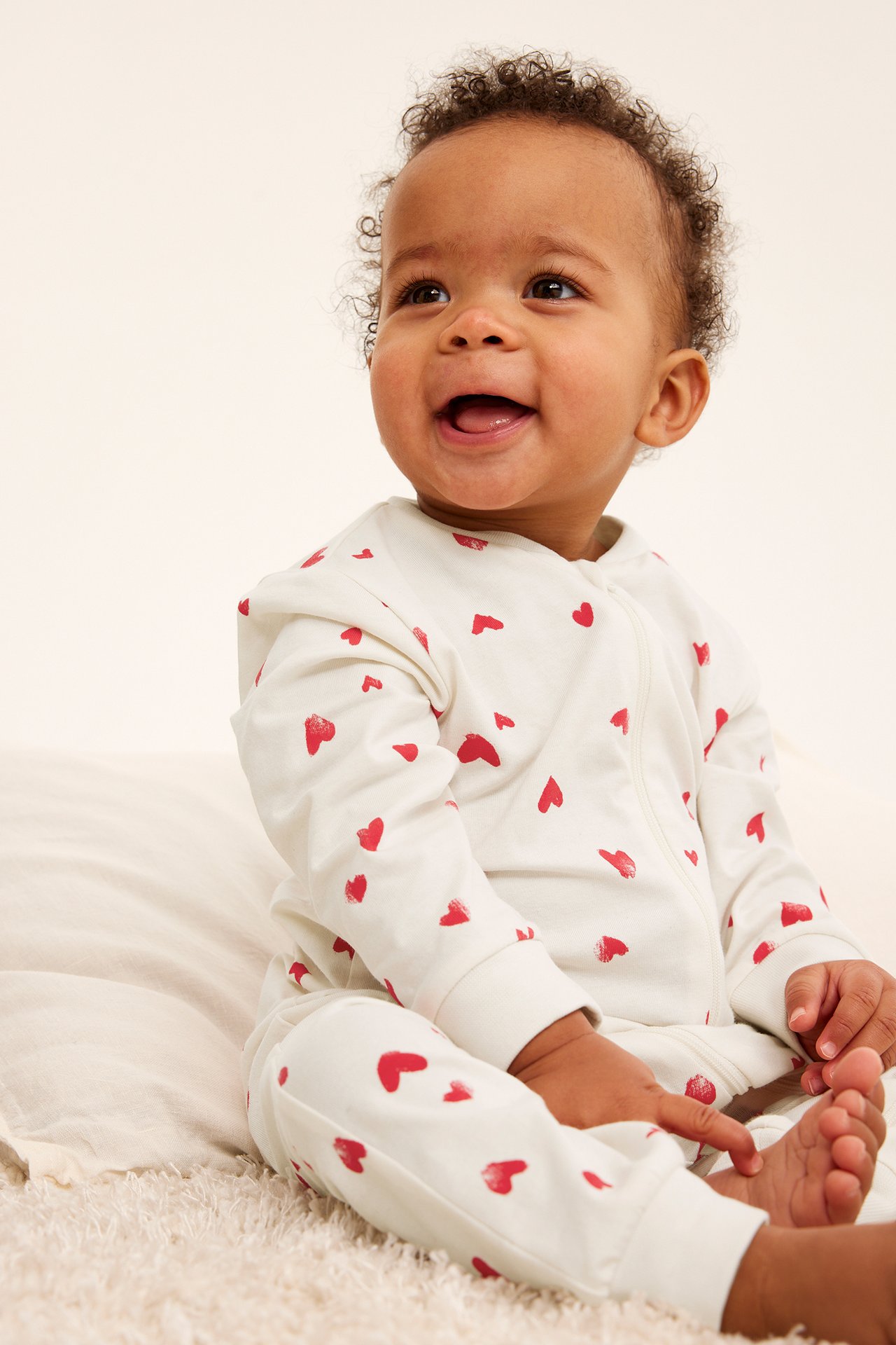 Forfra, en smilende baby i offwhite pysjamas med røde hjerter og glidelås foran.