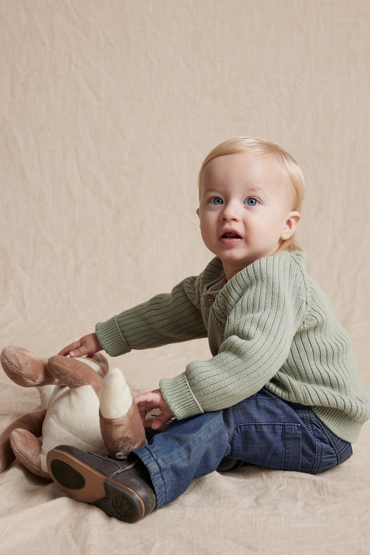 Baby i grønn strikket cardigan, blå jeans og mørke sko, leker med et brunt kosedyr.