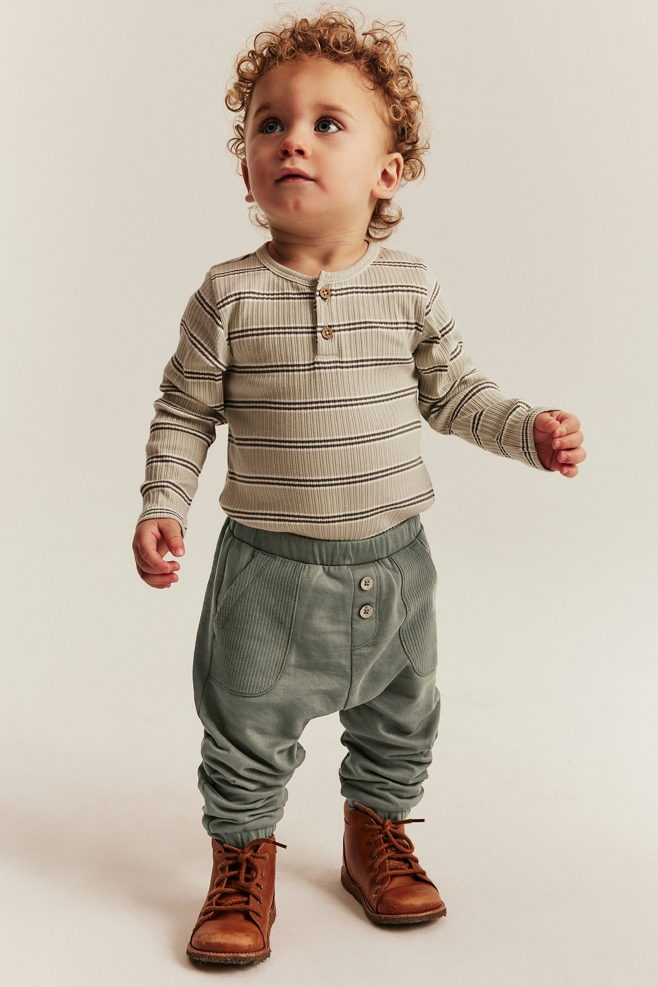 Framifrån: Baby i mörkgröna mjukisbyxor med knappar, randig topp och bruna kängor.
