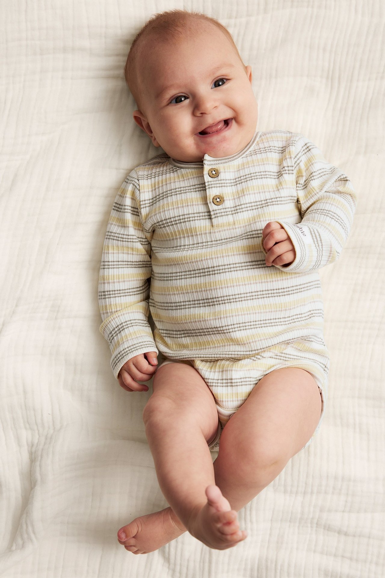 Hymyilevä vauva yllään luonnonvalkoinen, keltainen, harmaa raidallinen pitkähihainen puuvillabody napeilla, ylhäältä.
