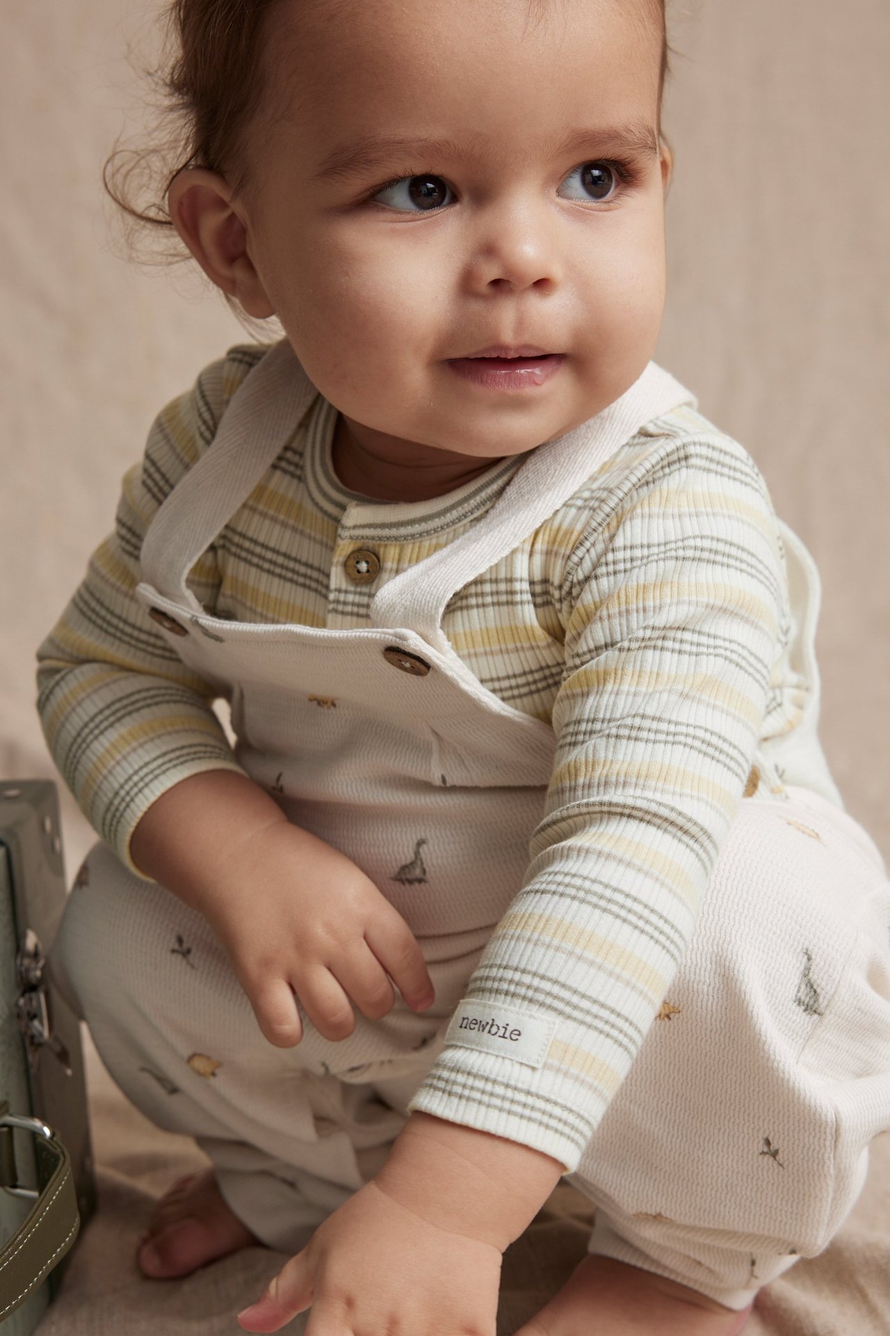 Lähikuva edestä vauvasta, jolla on raidallinen luonnonvalkoinen, keltainen ja harmaa body luonnonvalkoisten haalareiden alla.