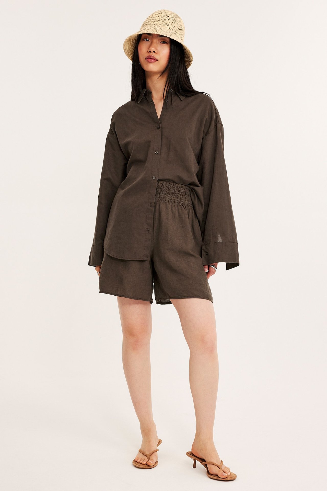Frontalvisning: Kvinne i mørkebrune linskjorts med smockmidje, matchende skjorte, beige stråhatt og brune sandaler.