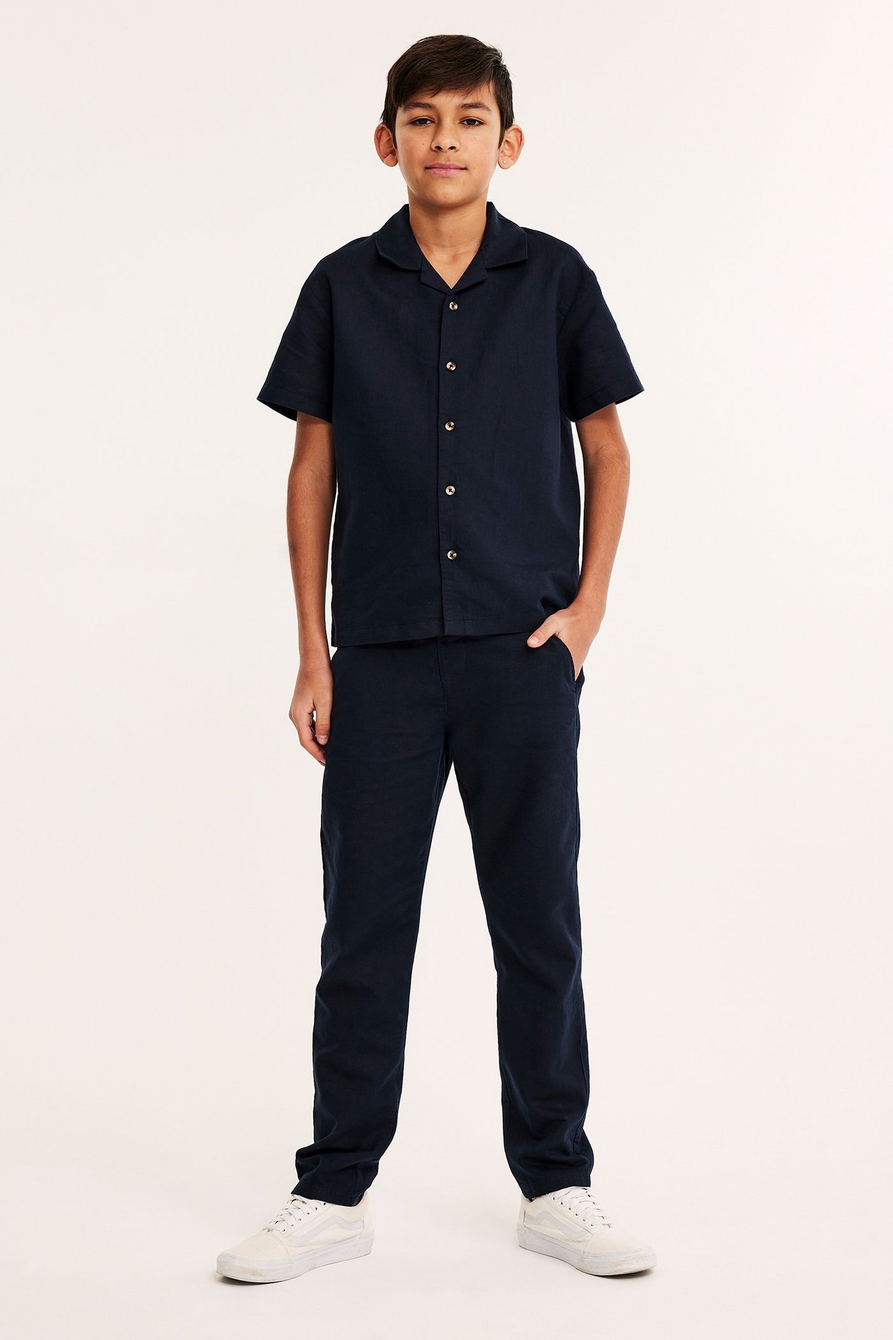 Widok z przodu dziecka w granatowych spodniach z mieszanki lnu, pasującej koszuli z krótkim rękawem i białych trampkach.