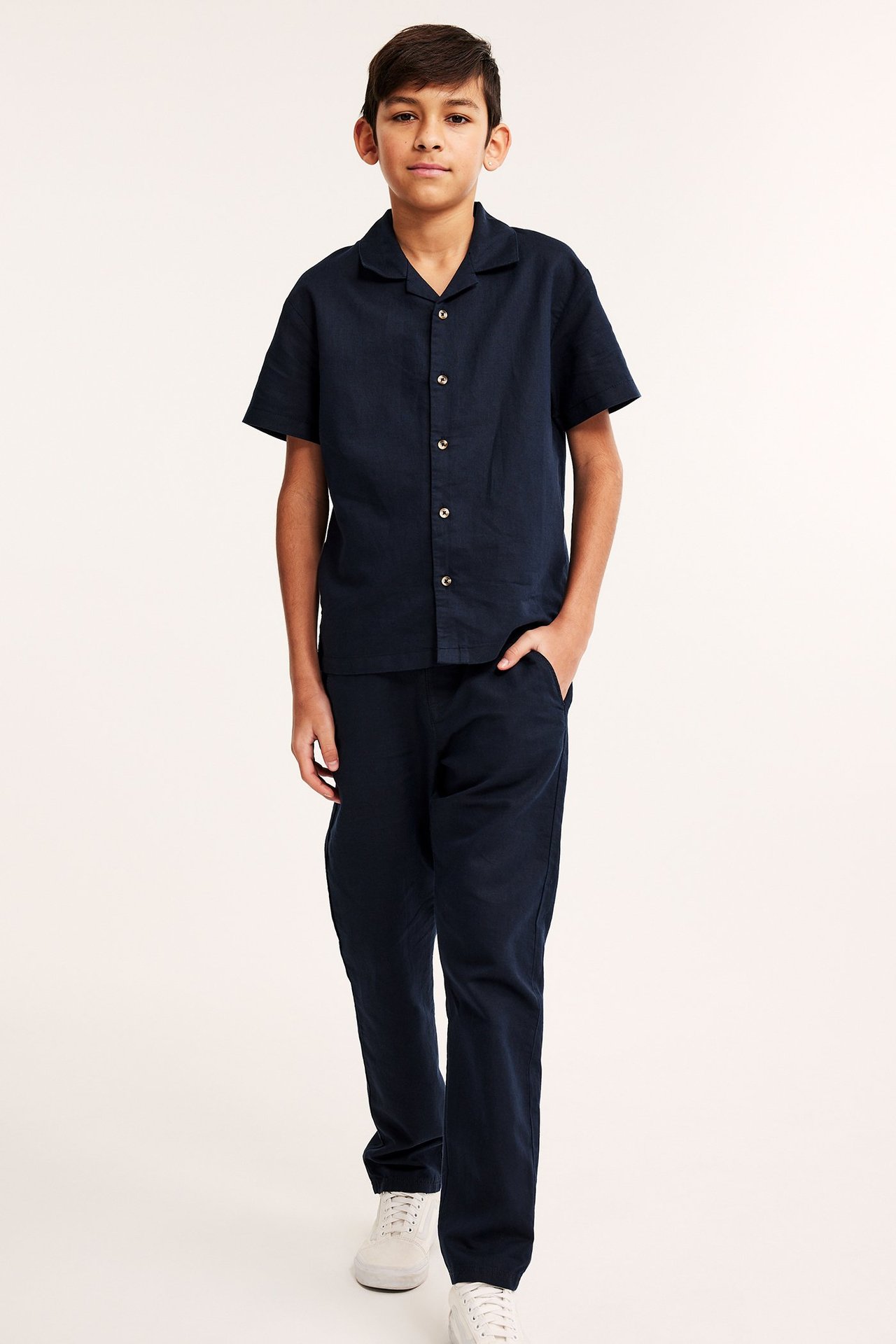 Widok z przodu dziecka w granatowych spodniach z mieszanki lnu, pasującej koszuli z krótkim rękawem i białych trampkach.