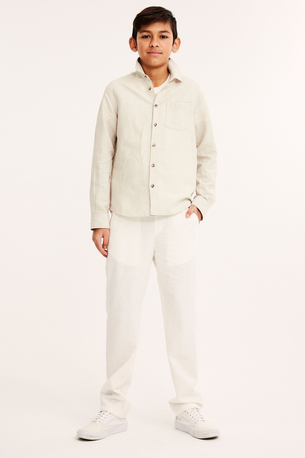 Framifrån: Barn i ljusbeige linneblandningsskjorta, vit t-shirt, vita byxor och vita sneakers.