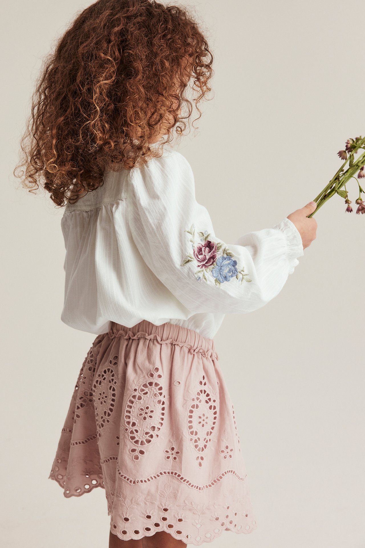 Barn, bakfra: offwhite bomullsbluse med blomstrete puffermer, rosa broderie anglaise-skjørt.