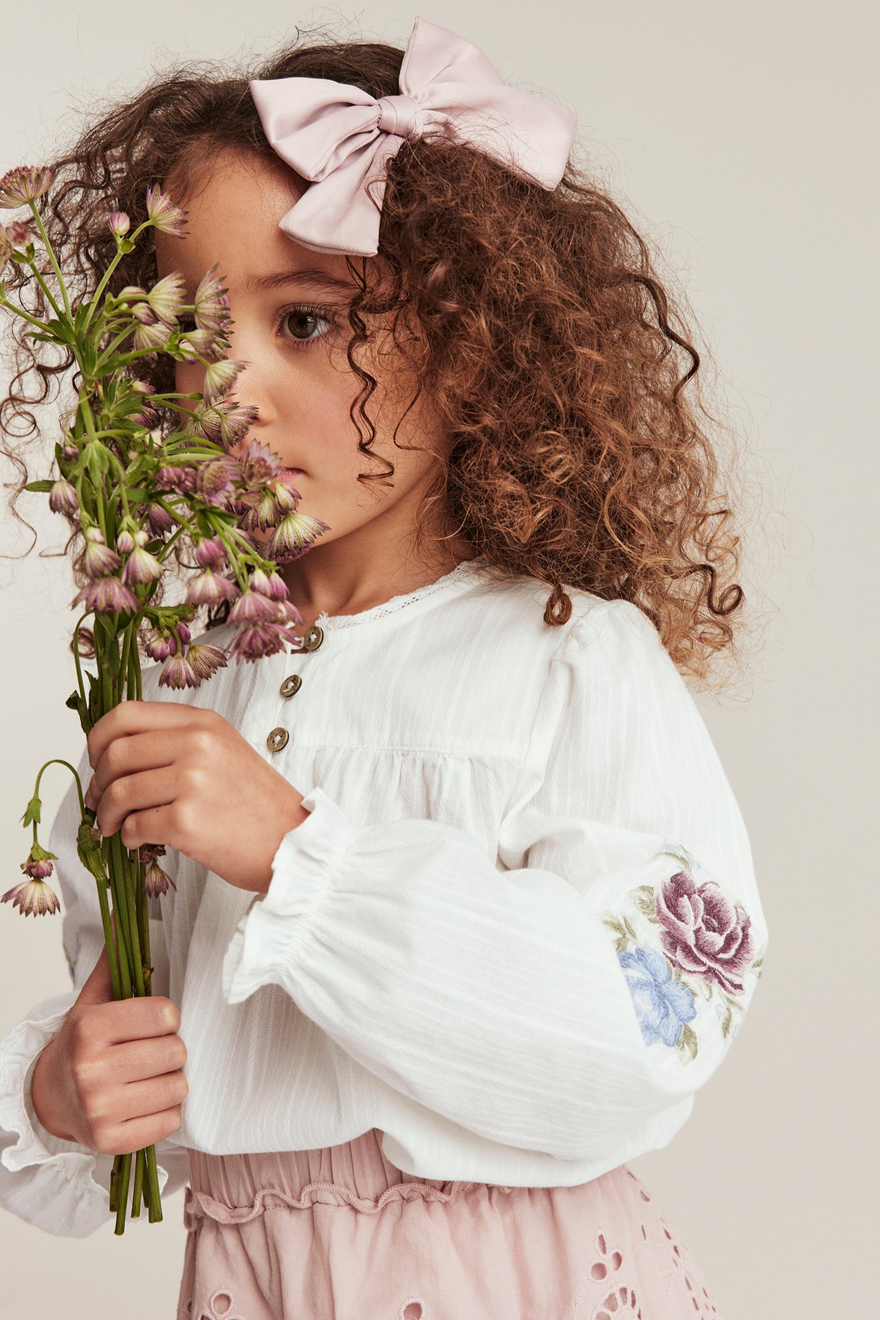 Barn i offwhite bluse med blomsterbroderte puffermer, rosa skjørt og rosa sløyfe, holder blomster.