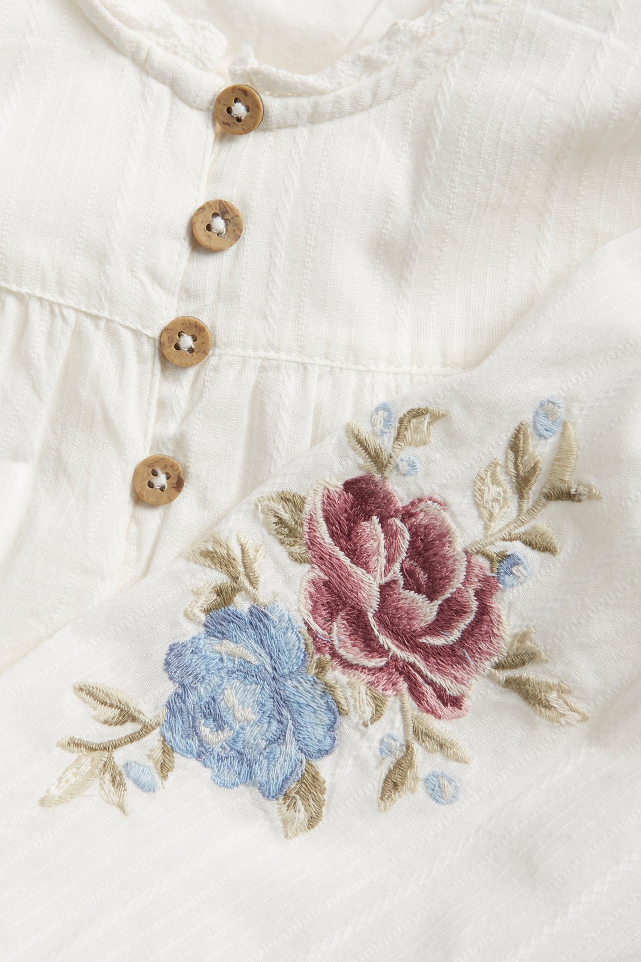 Nærbilde av en offwhite teksturert bomullsbluse med treknapper og rosa og blå blomsterbroderi.