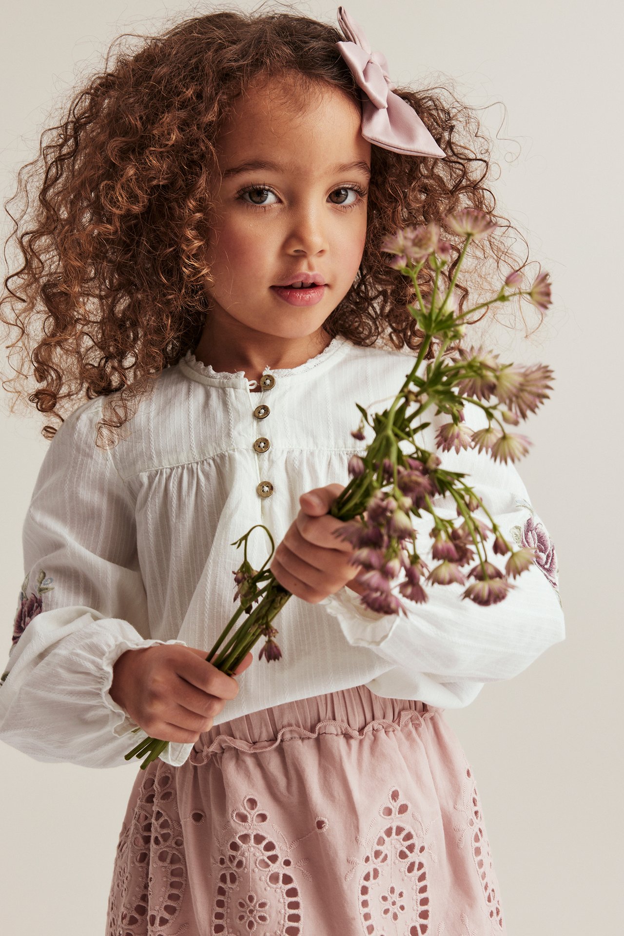 Frontvisning: Barn i offwhite brodert puffermebluse, rosa hullbrodert skjørt, rosa sløyfe, holder blomster.