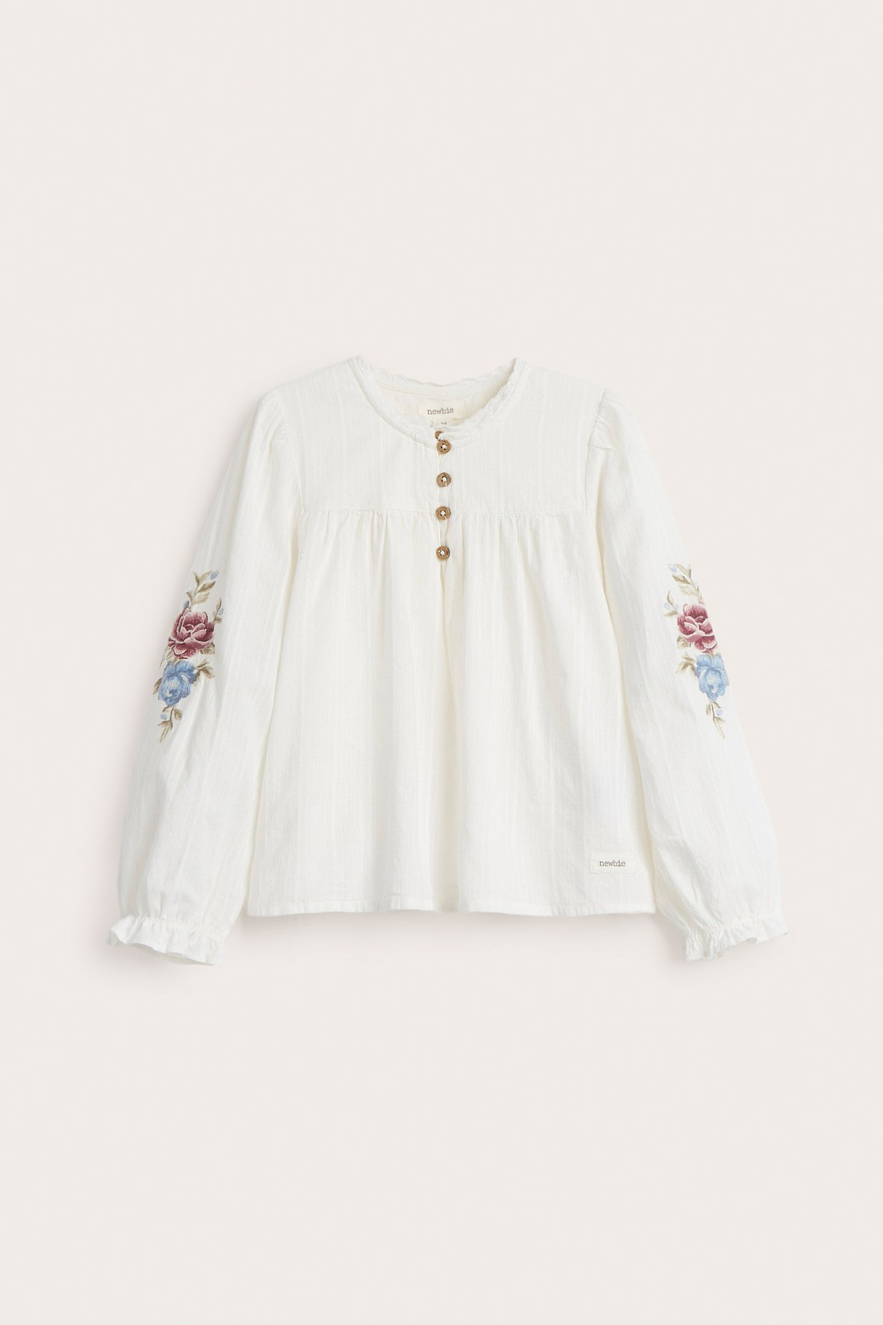Forfra: Offwhite barnebluse i strukturert stoff med blomsterbroderte puffermer og knappelukking foran.