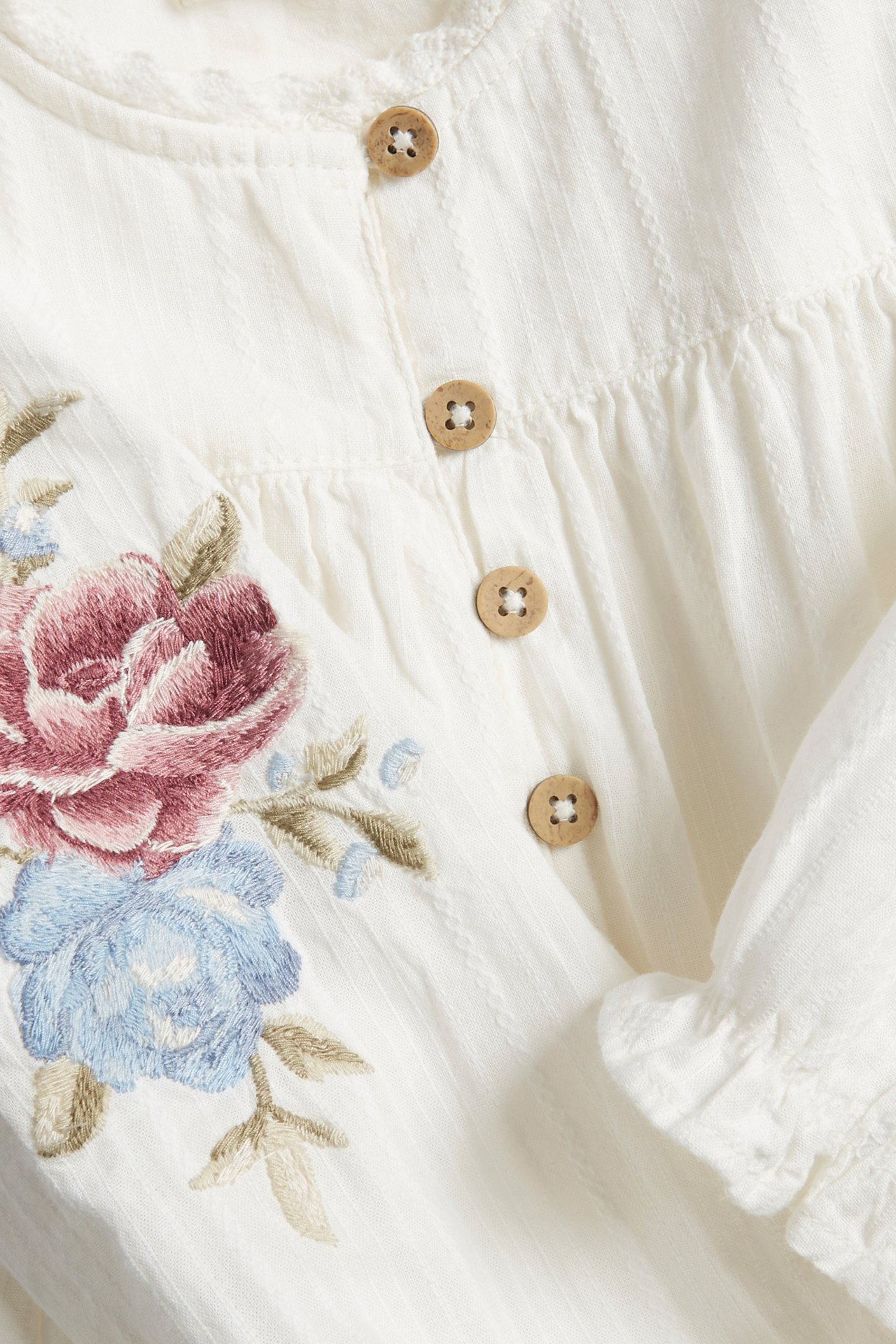 Närbild på en offwhite texturerad bomullsbody med blommig brodyr och träknappar fram.