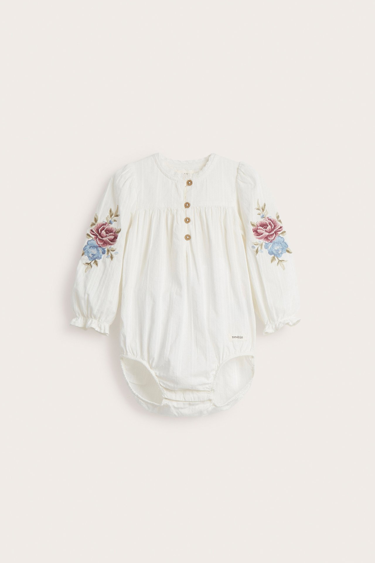 Framsida av en offwhite babybody i bomull med broderade puffärmar och knappar fram.
