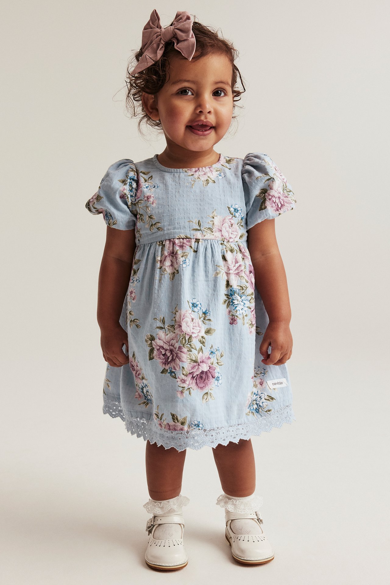 Framsida: Baby i blå blommig klänning med ballongärmar, spetskant, rosa rosett, vita strumpor, vita skor.