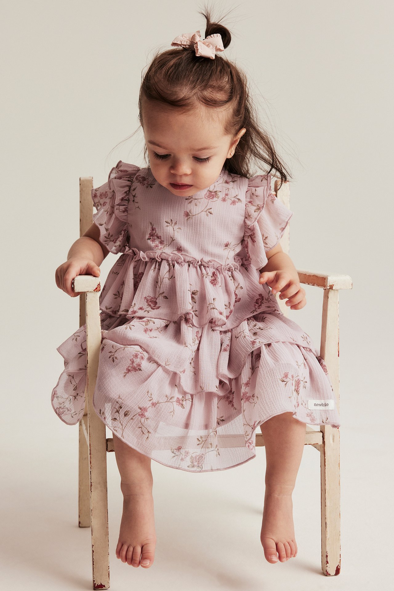 Forfra, en babyjente i rosa blomstret chiffonkjole med volanger, volangermer og rosa hårsløyfe.