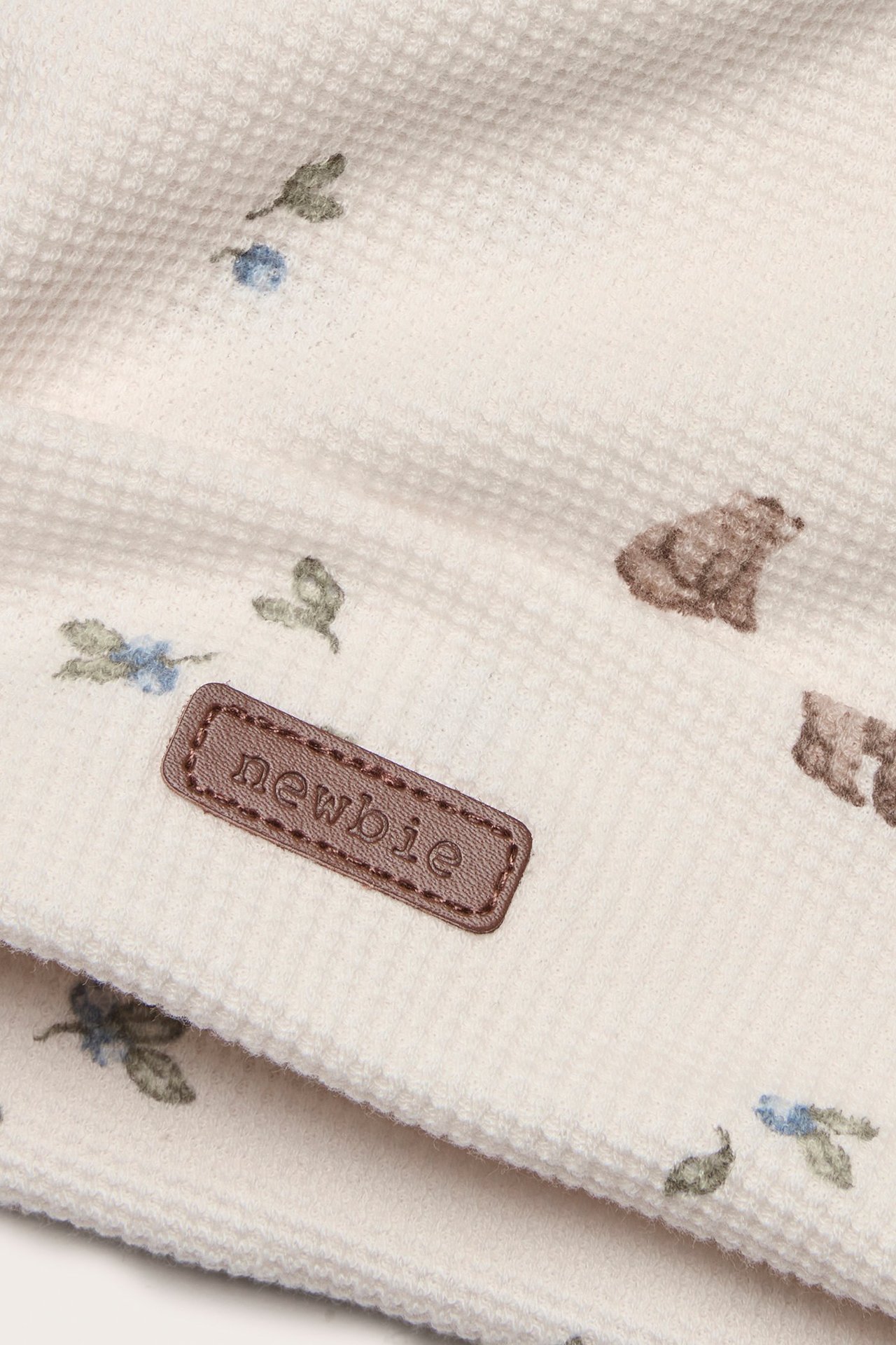 Nærbilde av en offwhite vaffelstrikket lue med bjørne- og bladmotiv og en brun 'newbie'-etikett.