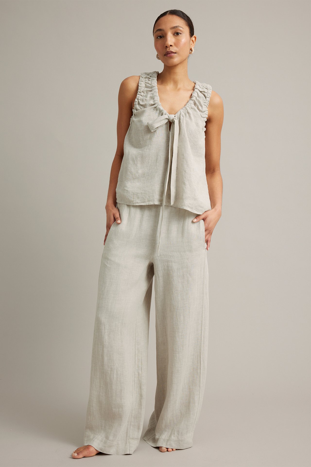 Frontvy: Kvinna i melerade beiga linnebyxor med vid passform och matchande volangtopp med knytband.