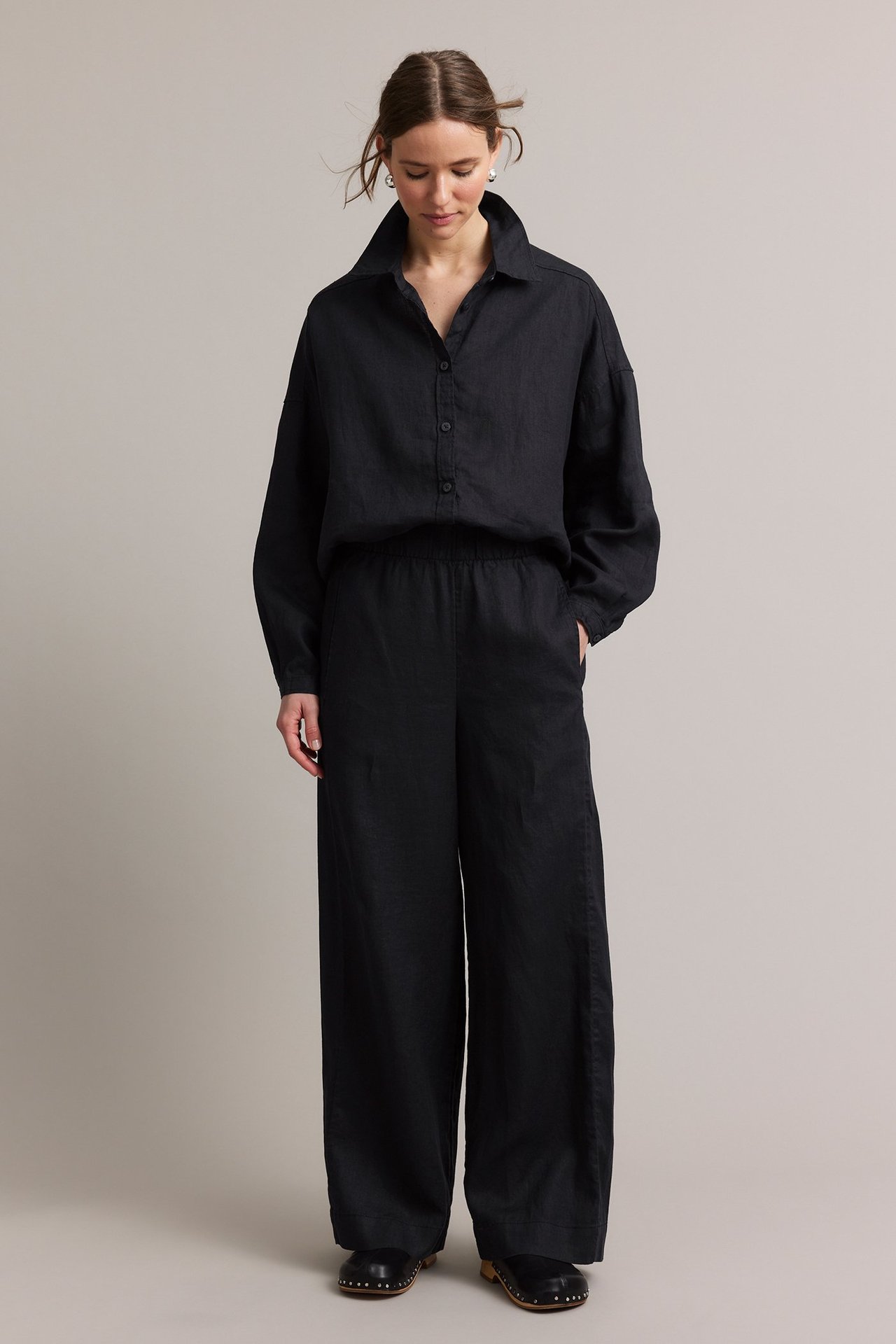 Naisen etunäkymä mustissa pellavahousuissa, joissa suorat lahkeet, yhteensopiva paita, mustat puukengät ja hopeakorvakorut.