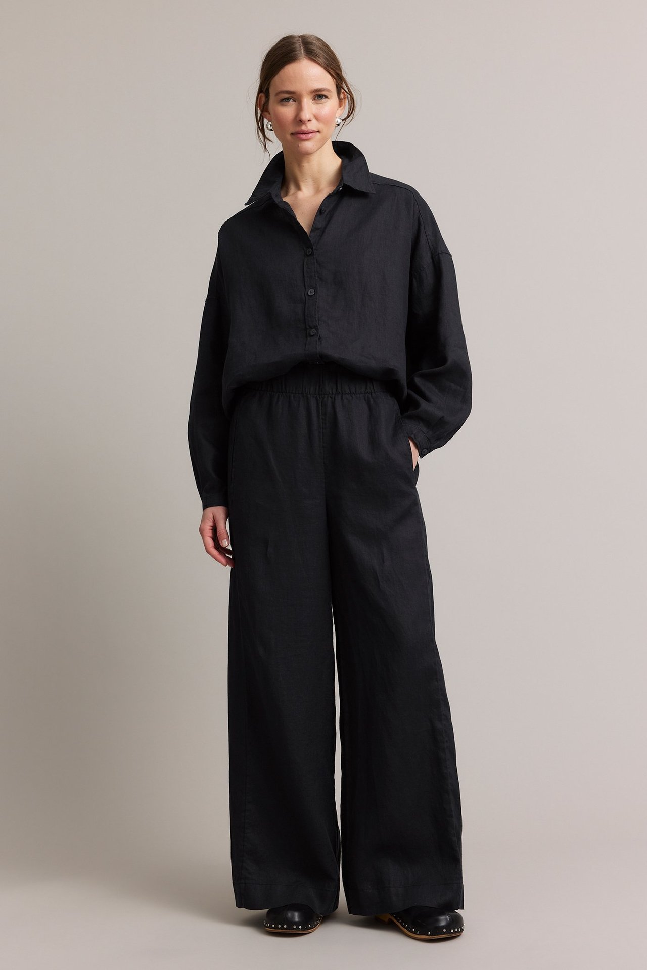 Nainen edestäpäin: mustat leveät pellavahousut, musta pellavapaita ja mustat puukengät.