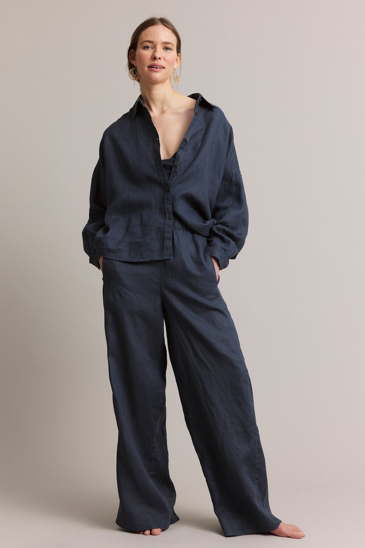 Helkroppsbilde av kvinne i mørkeblå rette linbukser, matchende linskjorte og gulløreringer.