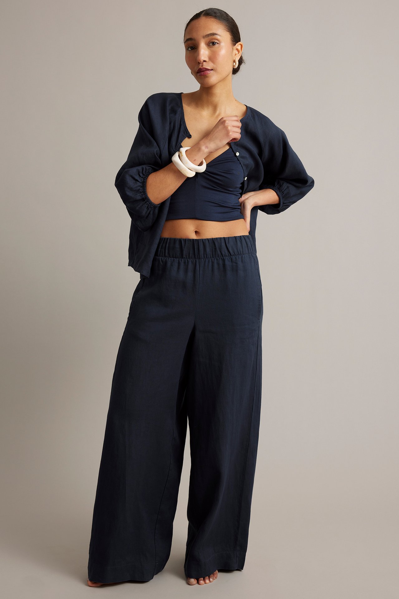 Frontvy: Kvinna i mörkblå linnebyxor med vida ben, matchande kofta, kort topp och ljusa armband.