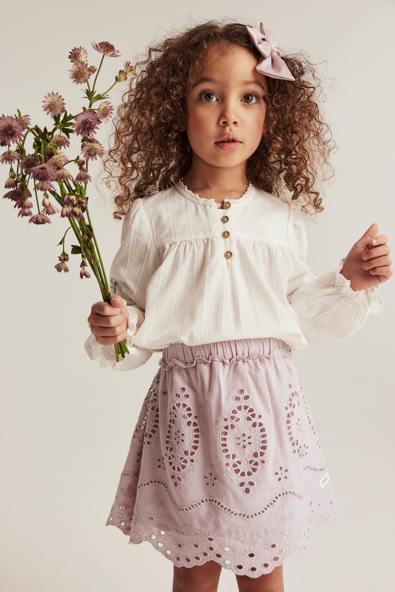 Framsida av ett barn i rosa broderad kjol, vit blus och rosa rosett, håller lila blommor.
