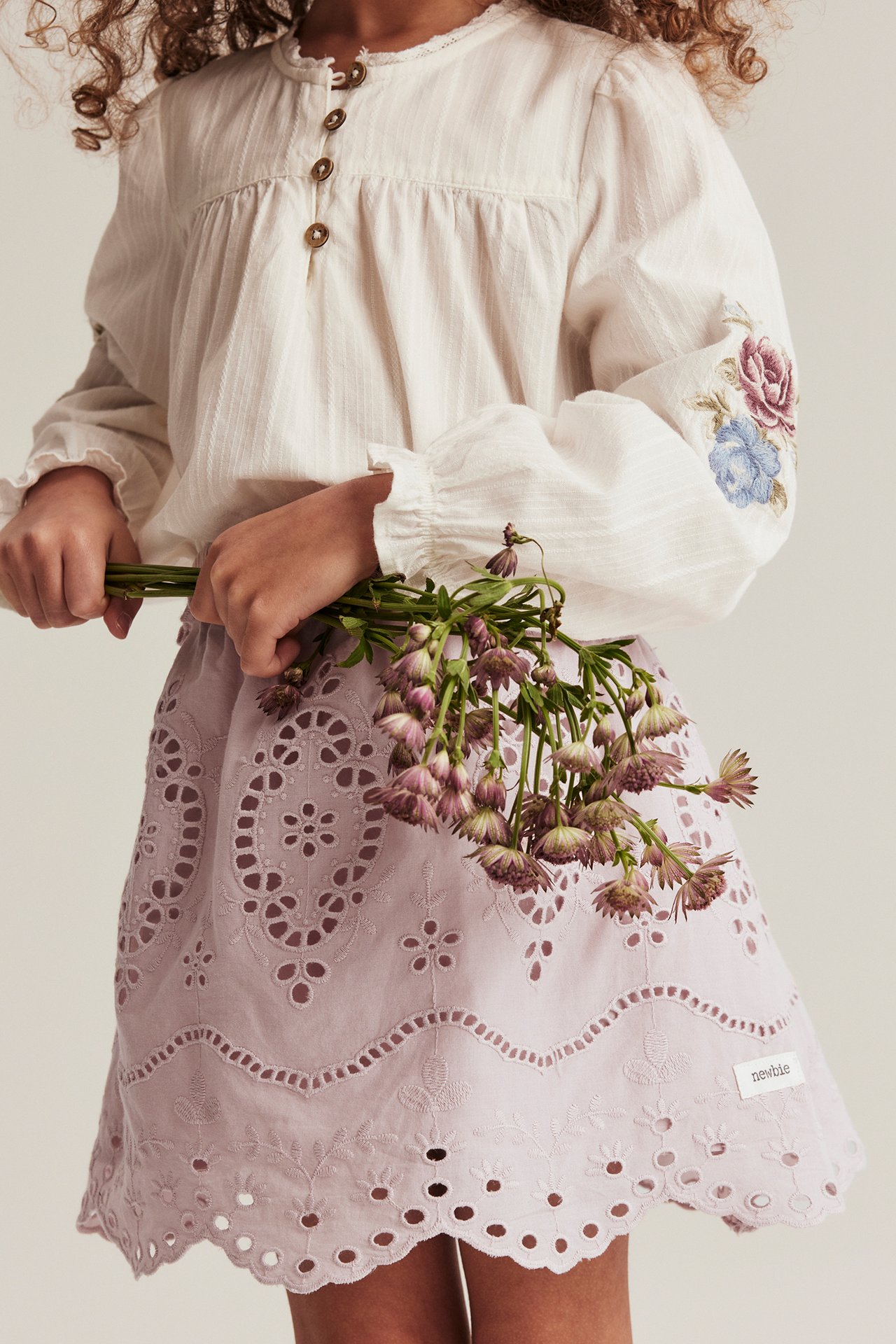 Framifrån: Kid i rosa broderad hålkjol och benvit blus med blommiga ärmar, håller blommor.